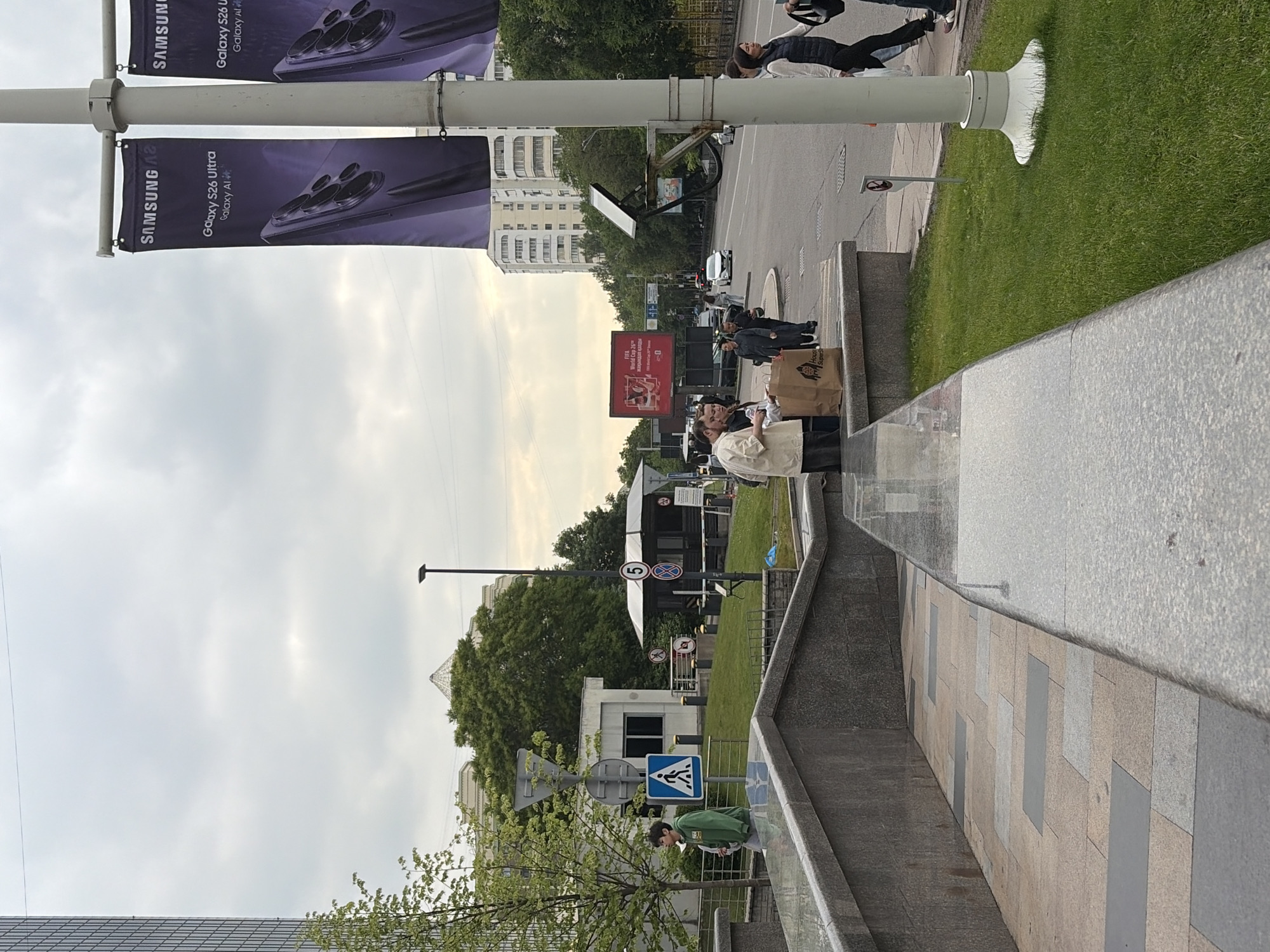 The image shows a street scene. A street light pole on the left displays two purple Samsung banners. Pedestrians are present on the sidewalk. A man is holding brown paper bags. A blue crosswalk sign is visible. Further down the street is a large red advertisement and various other street signs. The sky is overcast. A green lawn borders the walkway to the right. The street contains vehicle and pedestrian traffic. A tall building can be seen in the background.