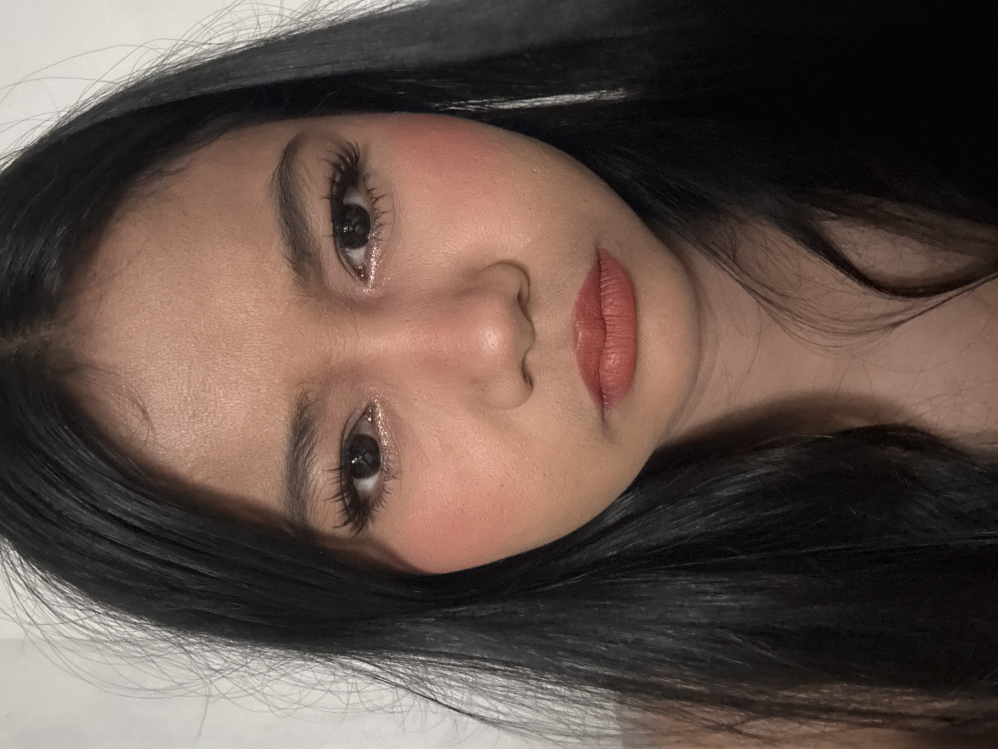 A young woman with black hair and makeup is looking directly at the camera. She is wearing blush on her cheeks and lipstick on her lips. Her hair is long and straight, and it frames her face. The background is a plain white wall. The lighting is soft and diffused, creating a gentle glow on her skin. The image appears to be a selfie, taken at close range, likely in an indoor setting. The woman is the sole subject in the image, and there are no other objects or people visible. The scene does not contain any specific indicators of a North Jakarta, Indonesia location.