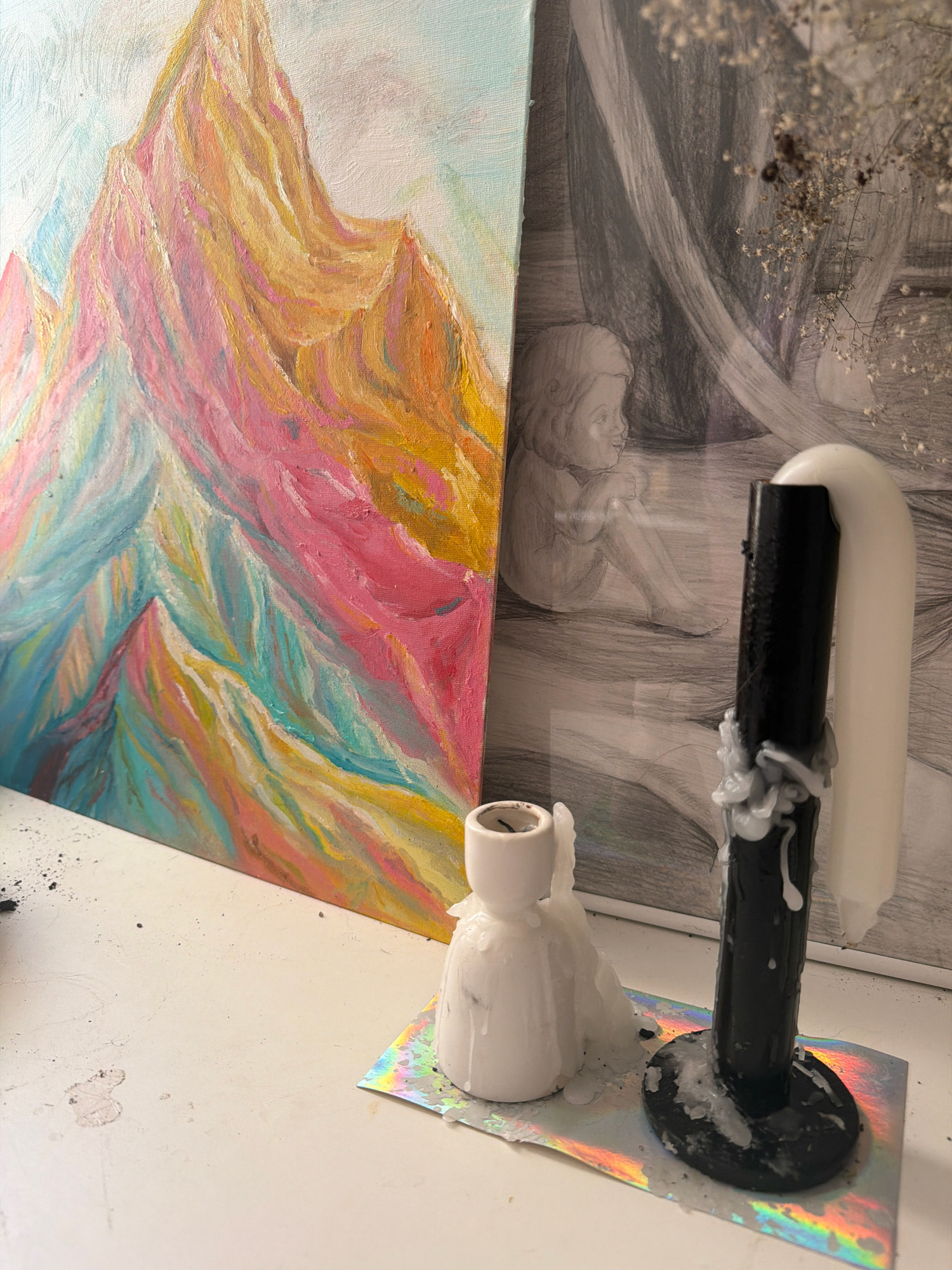 The image shows a still life arrangement on a white surface. In the foreground, there are two white candles with melted wax dripping down them. One candle is in a white holder, and the other is a thicker, black candle with white wax. Both are placed on a piece of iridescent holographic paper. Behind the candles, there are two pieces of art. To the left is a colorful, abstract painting of mountains with pink, yellow, and blue hues. To the right is a framed pencil sketch of a child sitting next to draped fabric. A few dried flowers are visible in the upper right corner. The scene appears to be indoors, possibly on a shelf or desk. There is no indication of the city of Samara, Russia, in the image.