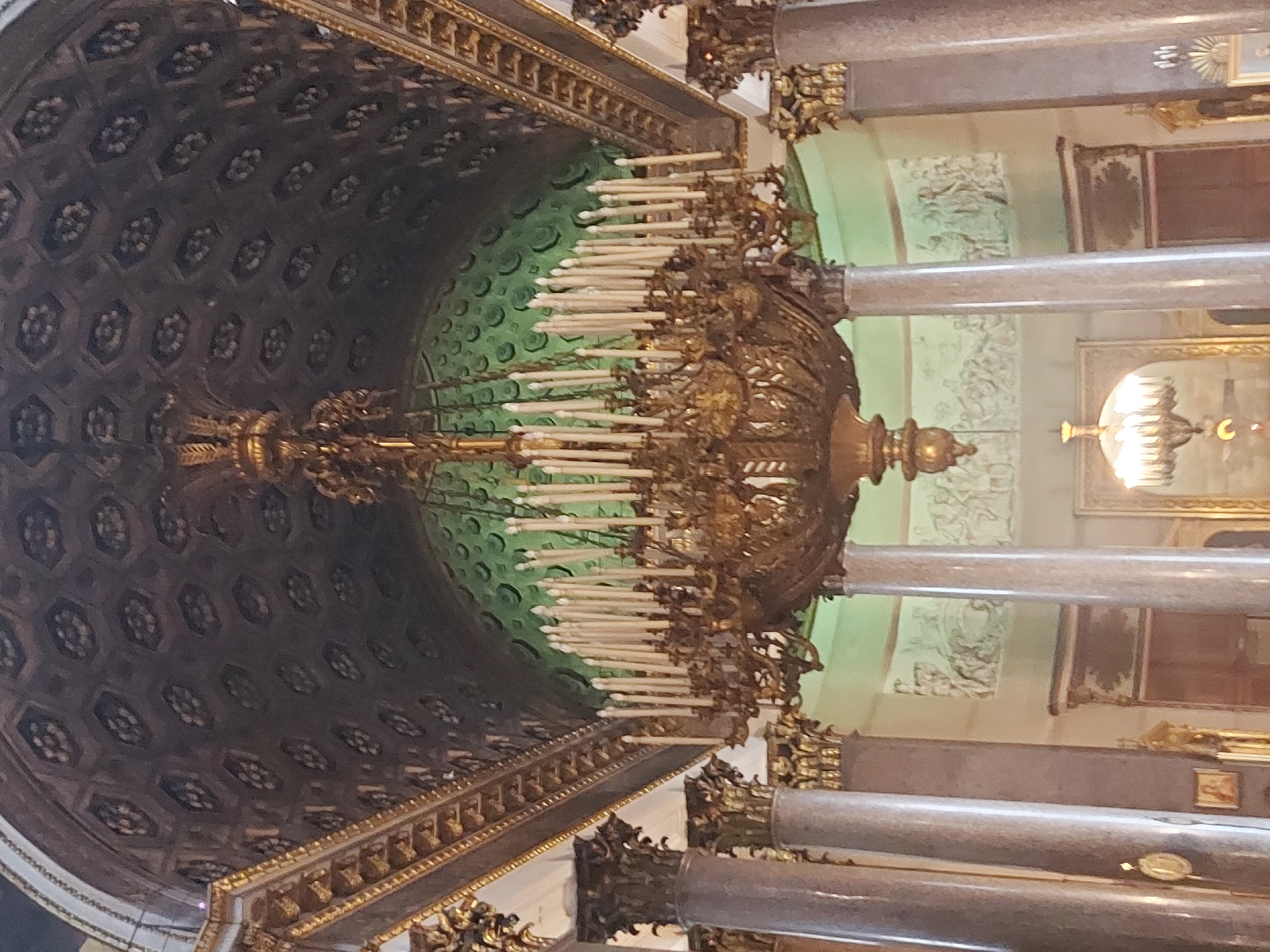 The image captures a fragment of the interior of a grand structure in an unknown location, potentially a cathedral or palace. The composition is oriented vertically, presenting an upward view towards an ornate ceiling and surrounding architectural details.
At the center of the frame is a massive, gilded chandelier, densely adorned with multiple tiers of candles. The chandelier's intricate framework is visible, suggesting high craftsmanship and historical significance.
The ceiling design features a pattern of dark, geometrically shaped panels, possibly coffers, interspersed with floral or rosette designs. The ceiling's perimeter is framed by elaborate, gilded molding.
To the right of the chandelier, tall, cylindrical marble columns are prominent. The walls behind these columns are decorated with pale green panels and reliefs. In the background, additional architectural elements, including what appears to be a framed artwork or mural, are partially visible.
The overall scene conveys a sense of opulence and grandeur, reflective of classical or baroque architectural styles. The lighting, predominantly from the chandelier and unseen sources, casts shadows and highlights, adding depth and texture to the scene.
