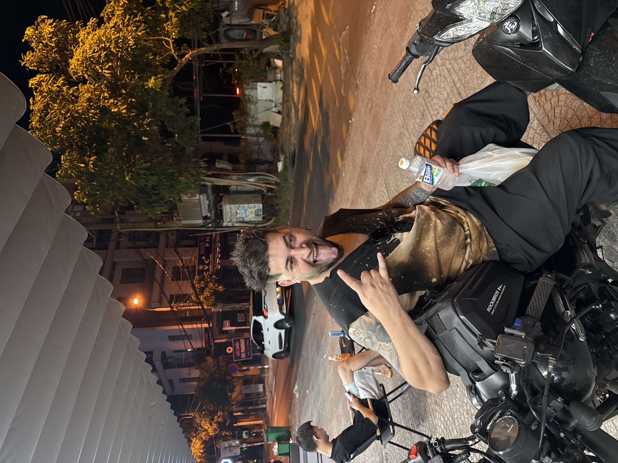A man is sitting on a motorcycle in an urban environment at night. The man has dark hair, a beard, and is wearing a black tank top and black pants. He has tattoos on his arms and is sticking his tongue out while making a "rock on" gesture with his hand. He is holding a water bottle and a white plastic bag. The motorcycle has a black ROCKBROS bag strapped to the front. Another person is sitting in a chair nearby, looking at a phone. A white car is parked on the street. The background shows buildings, trees, street lights, and commercial signs, including one for "CVC Pharmacy," indicative of a location in Vietnam. The photo shows that the scene is from a street in Đà Nẵng, Vietnam.