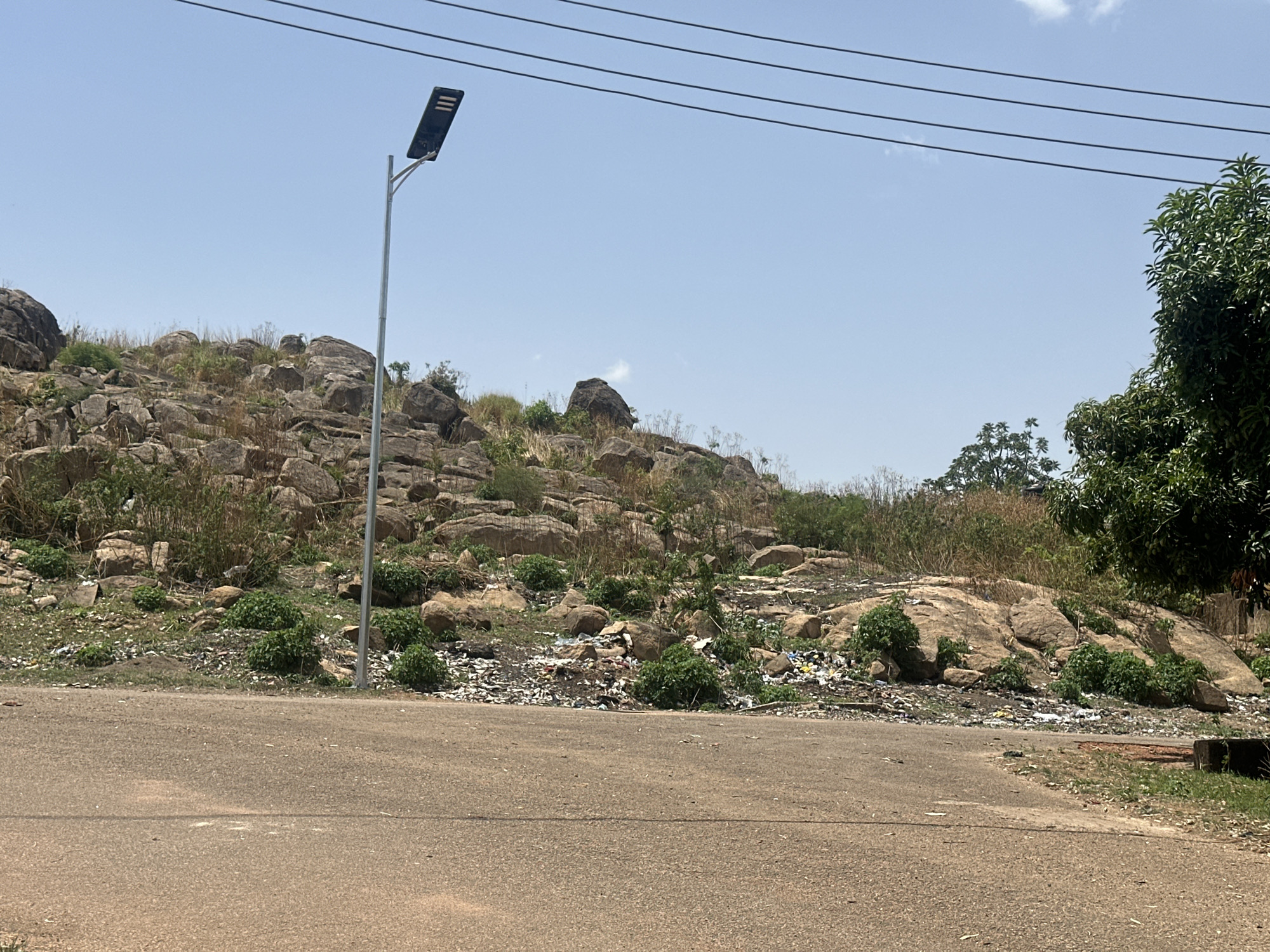 The image shows a roadside view in Jos, Nigeria. The road is paved and appears to be lightly traveled. A rocky outcrop rises immediately behind the road, covered with sparse vegetation, including small bushes and dry grasses. Significant amounts of litter are scattered along the base of the rocks, extending onto the roadside. A modern street light stands near the roadside. Overhead, several parallel power lines run across the top of the frame. A large green tree stands to the right. The sky is pale blue, with a few wispy clouds.