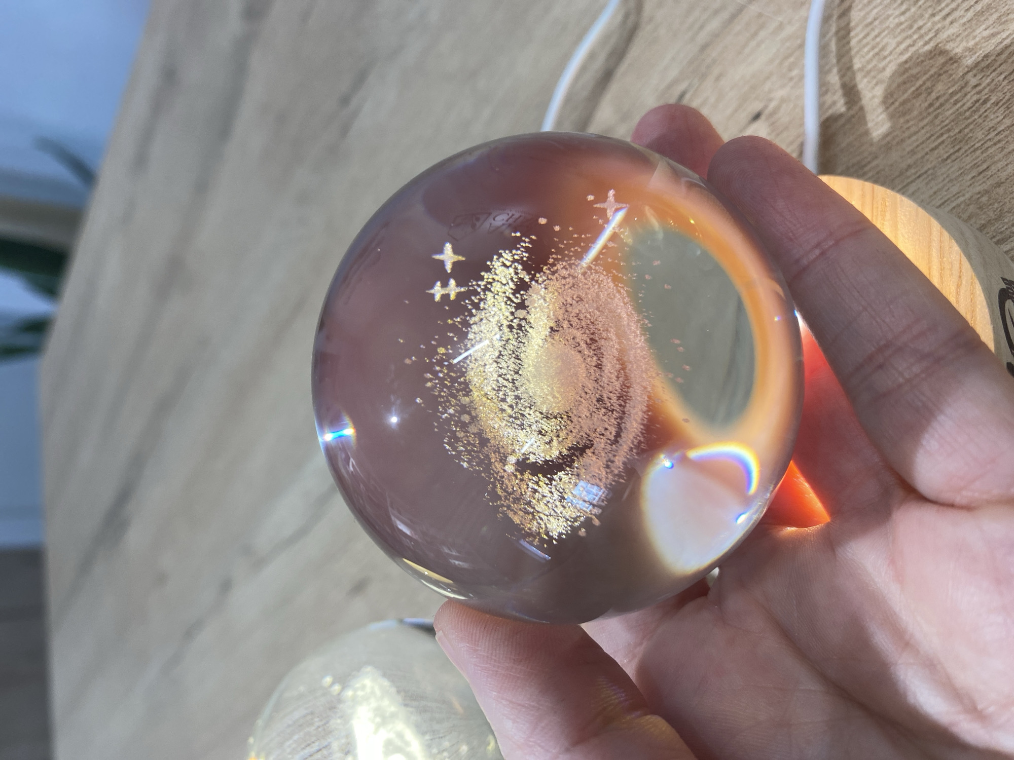 A hand holds a glass ball containing a glittering, irregular shape within. The ball is clear but tinted with a faint brownish hue on top, and it rests on a light-colored wooden base visible near the fingers. There is a white cable extending upwards from the base and out of the frame. The background features a light-colored wooden surface. The image does not contain information that can be used to determine the location in the city of Chaikovsky, Russia.