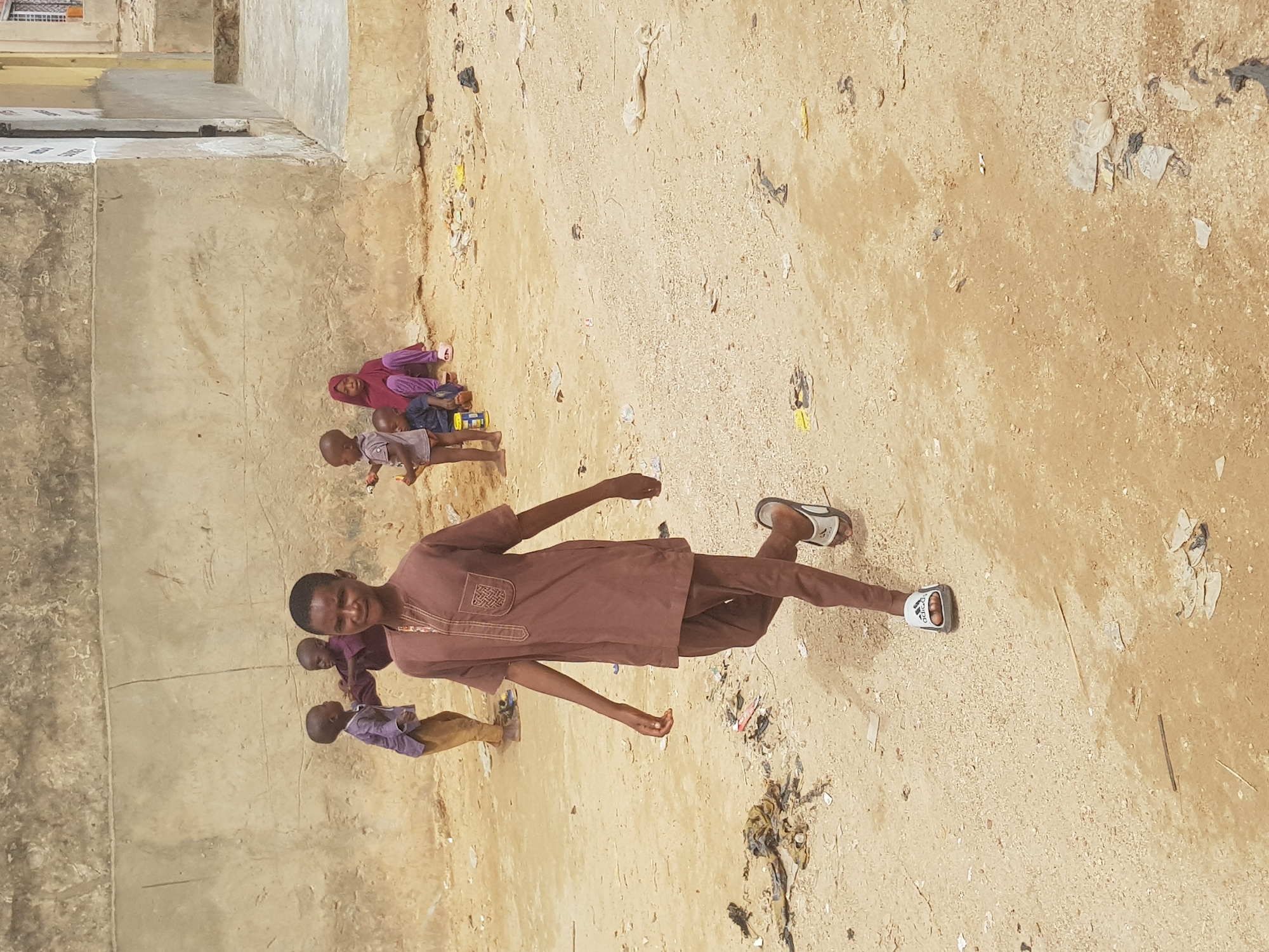 A brown-skinned man wearing a brown shirt and pants, with sandals, is standing in a dirt yard with trash scattered about. Three children stand behind him, near a cement wall. Two of the children are wearing purple and one is wearing pink. The location appears to be Dutsinma A, Nigeria.