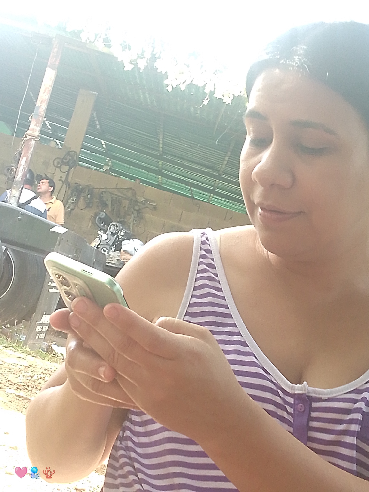 The image is a selfie taken outdoors, with the focus on a woman holding a light-green smartphone. She is wearing a purple and white striped tank top and her eyes are closed. In the background, a man in a yellow shirt is visible, looking up. The environment appears to be a workshop or garage, with various tools and equipment in the background. The lighting suggests it is daytime, possibly with bright sunlight. There are no clear indications of the specific location within Santa Ana, Venezuela.