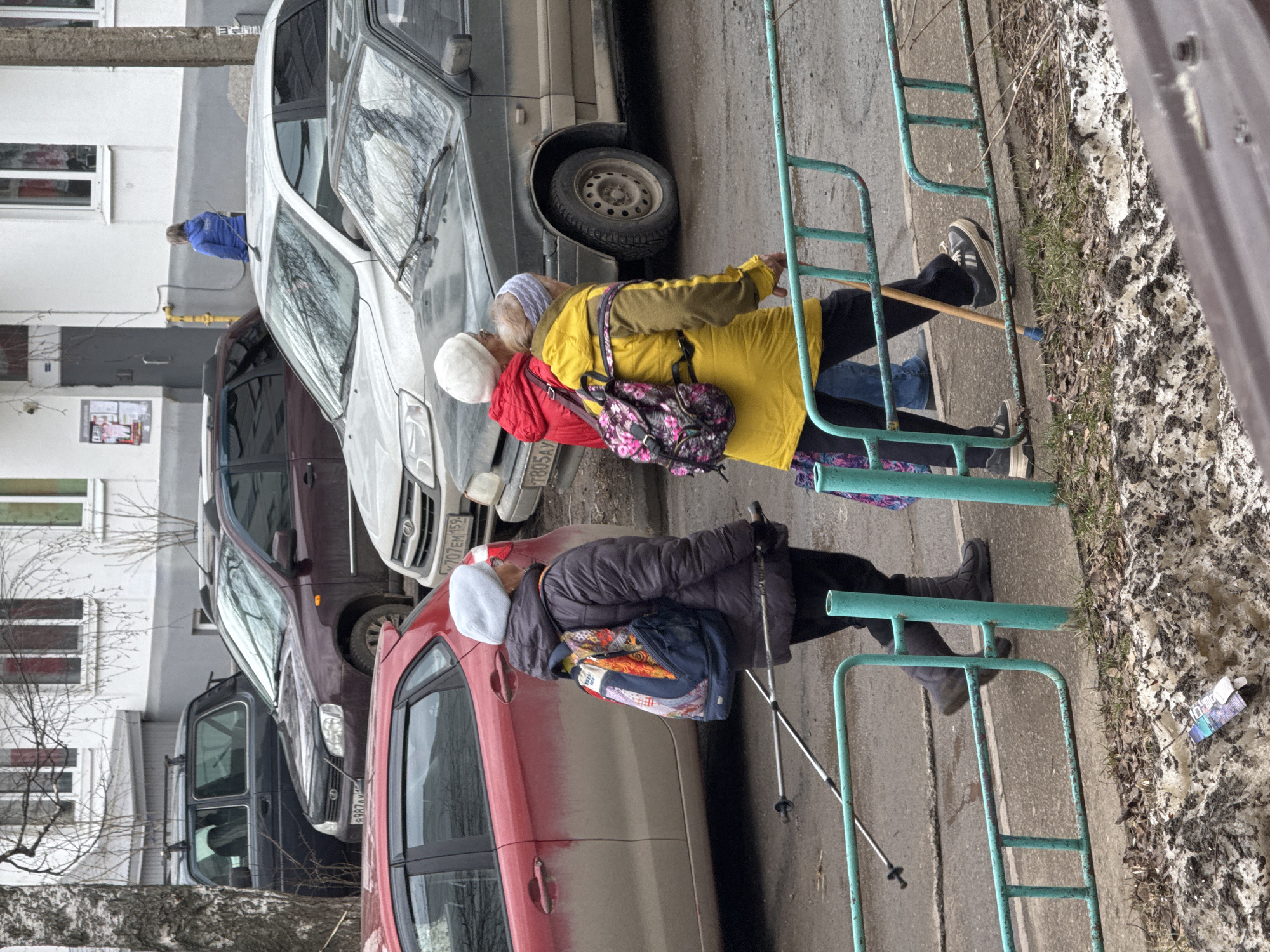 The image shows two elderly women in outerwear walking along a sidewalk next to parked cars. The woman closest to the camera wears a dark coat and a white hat, and carries a hiking pole. The second woman wears a yellow jacket over a red garment, with a floral backpack. Both women are using the railing of a green playground structure for support. The license plates on the cars suggest the location is in Russia. The environment includes bare trees, patches of snow, and a multi-story building. Based on the license plates, the location is most likely in the city of Chaikovsky, Russia.