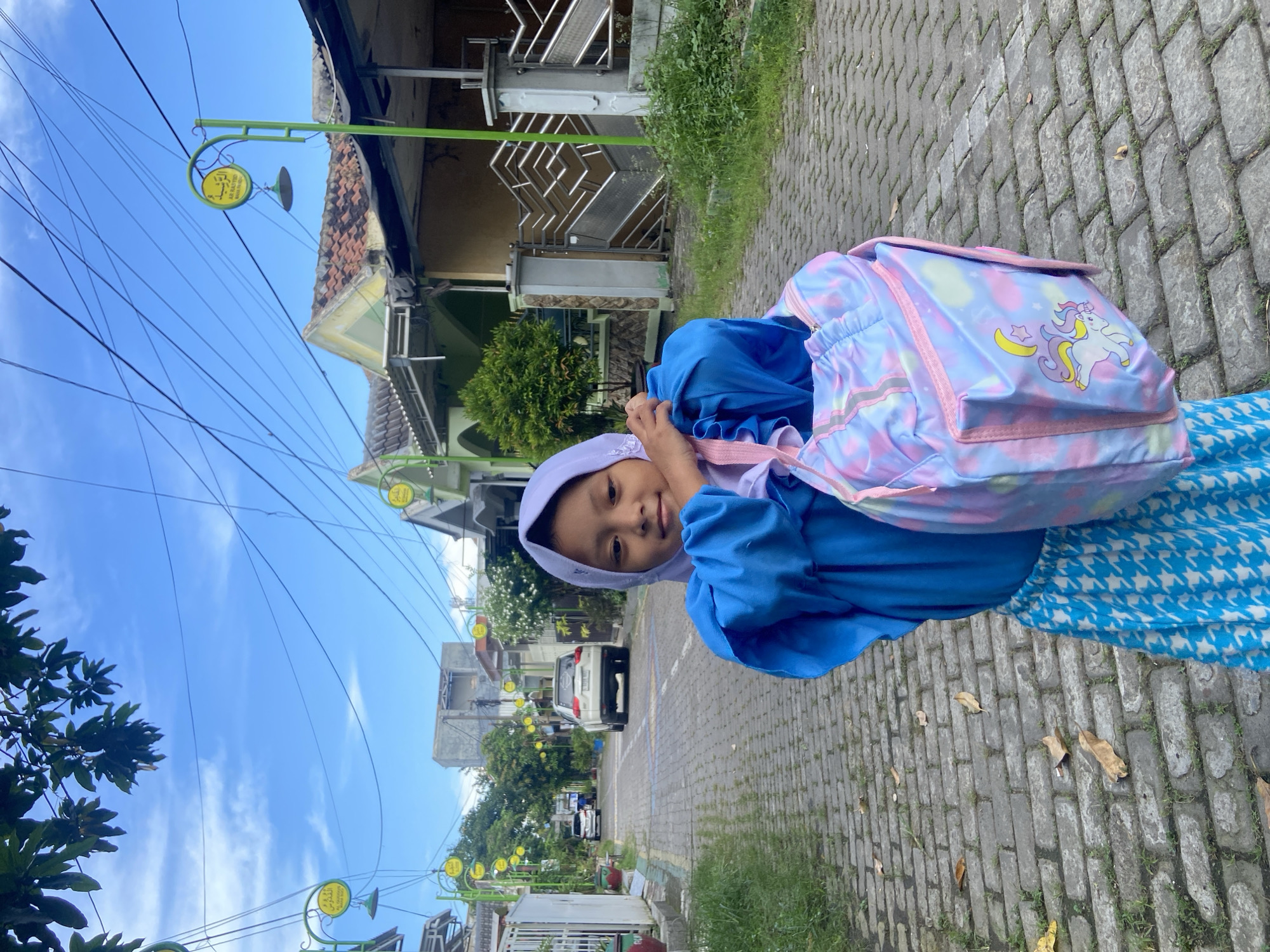 A young child, likely a girl, stands on a paved sidewalk in a residential area of Gelam, Indonesia. She looks directly at the camera with a gentle smile, her hands near her face, possibly adjusting her headscarf. She wears a light purple hijab and a vibrant blue dress or tunic over patterned blue and white lower garments. A colorful, light blue and pink tie-dye backpack, adorned with a cartoon unicorn, crescent moon, and star design, is on her back.
The scene unfolds under a bright blue sky with sparse white clouds, indicating clear daytime weather. Numerous black power lines crisscross the sky. The background features a street lined with residential buildings, some showcasing traditional tiled roofs. Along the sidewalk, green poles support circular yellow signs with Arabic script, one of which partially displays "Al-Hidayah" and "Ahlan wa Sahlan" (Welcome), suggesting local landmarks or institutions. Green foliage and trees are visible, and dried leaves are scattered on the grey brick pavers of the sidewalk. A white car is parked further down the street. The child's attire and backpack suggest she might be on her way to or from school. The image is captured at a Dutch angle, giving it a tilted perspective.