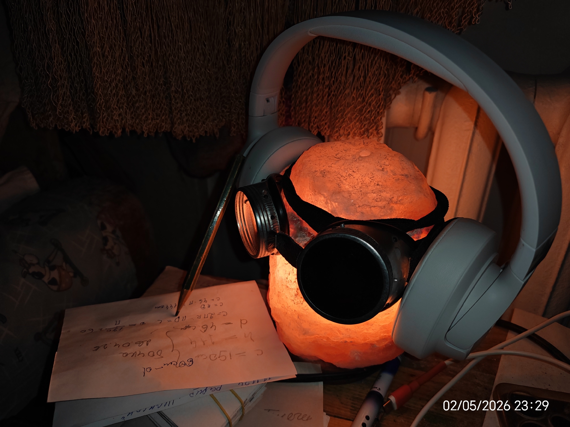 Here is a forensic description of the image:
The image shows a dimly lit scene with several objects arranged on a surface. Dominating the view is a pair of light gray wireless headphones placed around a glowing orange salt lamp. The lamp has a pair of steampunk-style goggles strapped around it. To the left of the lamp, there is a stack of papers with handwritten text and mathematical formulas. A pencil rests on top of the papers. A portion of a patterned pillow is visible on the left side of the scene, and some cables and pens are visible to the right, adding to the cluttered feel of the environment. The date "02/05/2026 23:29" is stamped on the lower right corner of the image, likely indicating when the photo was taken.