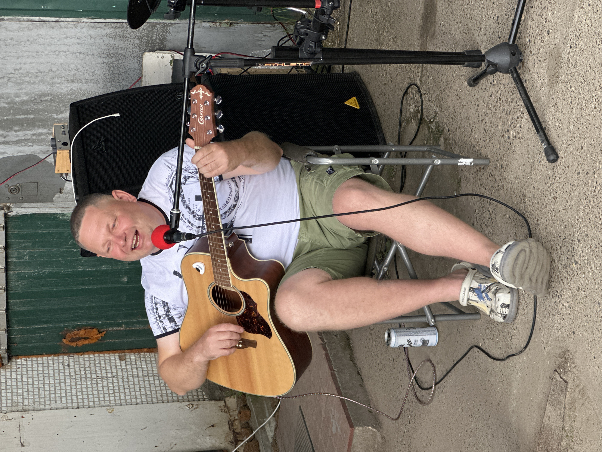An outdoor scene in Chaikovsky, Russia, features a middle-aged man sitting on a folding chair and playing an acoustic guitar. He is wearing a white t-shirt with a black and white pattern, green shorts, and colorful clogs. A red microphone is positioned in front of him, and a speaker is visible behind him. A can of drink rests on the ground near his feet. The background shows a tiled wall and a green metal door.