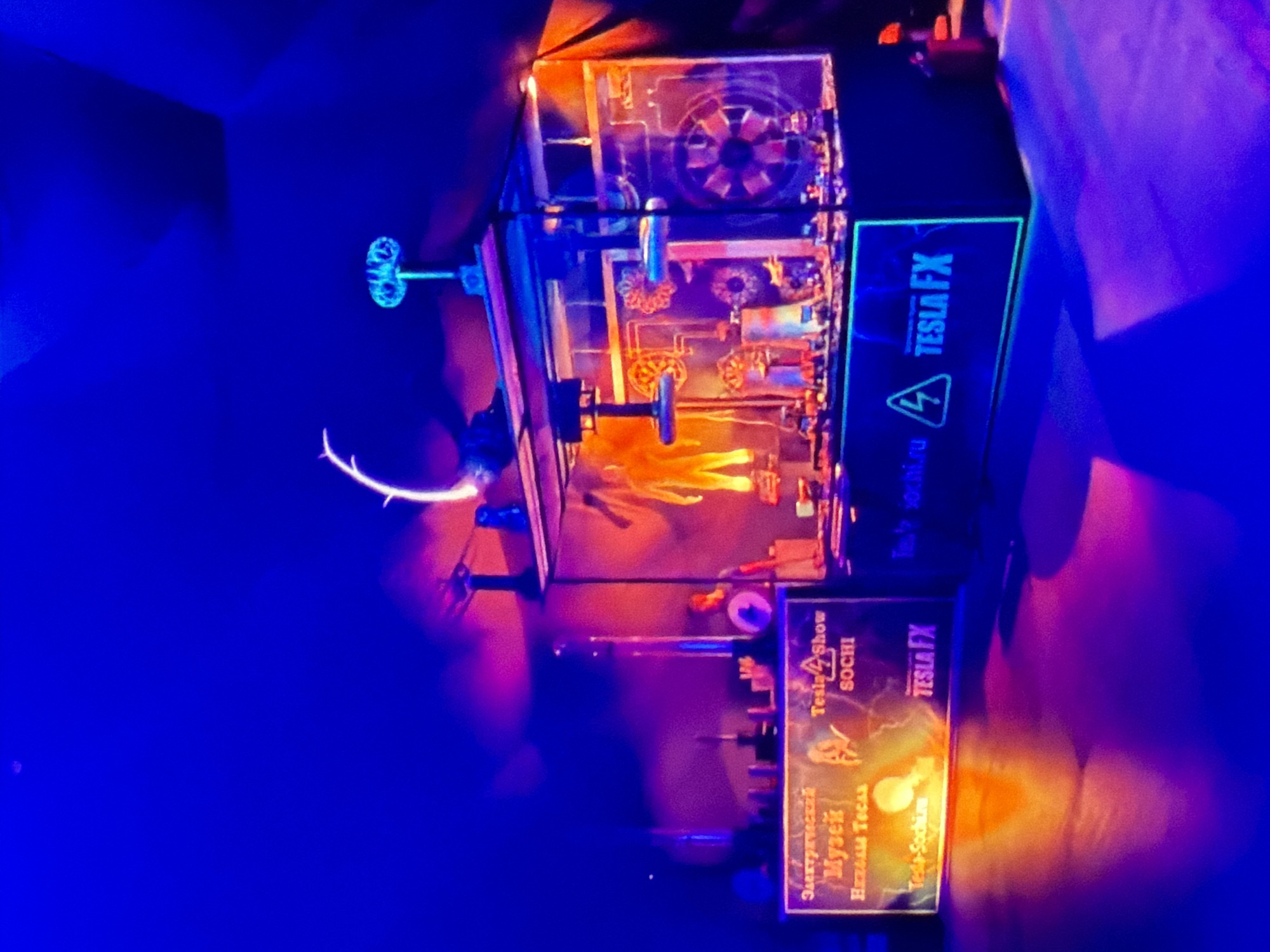 Here is a description of the image:
The image shows an illuminated exhibit, possibly at a Tesla-themed museum or attraction in Sochi, Russia. The exhibit features a complex arrangement of gears, pipes, and other mechanical elements encased in a transparent structure. The "Tesla FX" logo and website (tesla-sochi.ru) are visible on the side of the exhibit. Another sign reads "Electrical Museum of Nikola Tesla, Tesla Show Sochi". The scene is brightly lit with blue and orange/yellow hues, creating a dramatic effect.