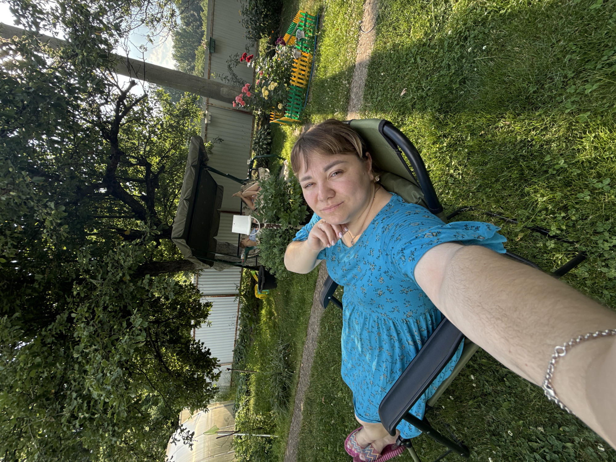 A woman is sitting in a lawn chair in a yard. She is wearing a blue floral dress and pink flip-flops. Her right arm is resting on the armrest of the chair, and she is looking at the camera. The background shows a fence, a swing set, a rose bush, and a greenhouse. The sun is shining, indicating it is daytime. The scene appears to be a backyard in a rural setting. There is no visible text in the image.