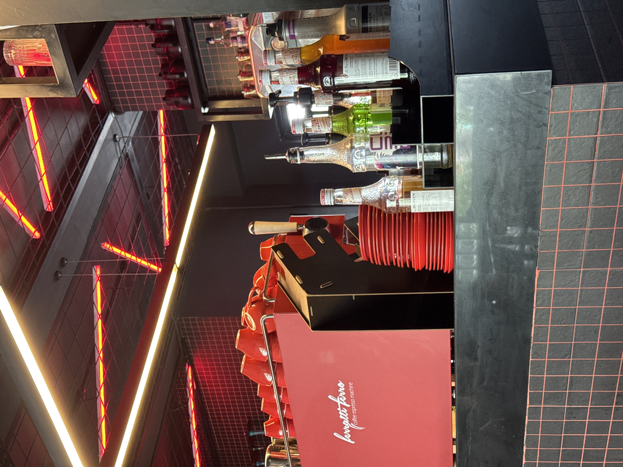 The image depicts a contemporary coffee shop counter area, likely in Kuta Selatan, Indonesia, characterized by a sleek, modern design with a focus on dark materials and vibrant red accents. Dominating the left side is a bright red espresso machine with the "ferrari ferro coffee espresso machine" logo clearly visible. A row of matching red coffee cups is stacked on top of the machine. To the right of the espresso machine, the counter is lined with various bottles of syrups, mixers, and spirits, many of which are Monin brand. These bottles are arranged on shelves and within a dispenser unit. In the background, the ceiling features a grid of dark tiles interspersed with bright red and yellow neon lighting fixtures. The counter itself is made of a dark, reflective material, and below it, a wall tiled with small, dark squares and thin red grout lines is visible. The overall impression is a well-lit, stylish, and functional space designed for coffee preparation and service.