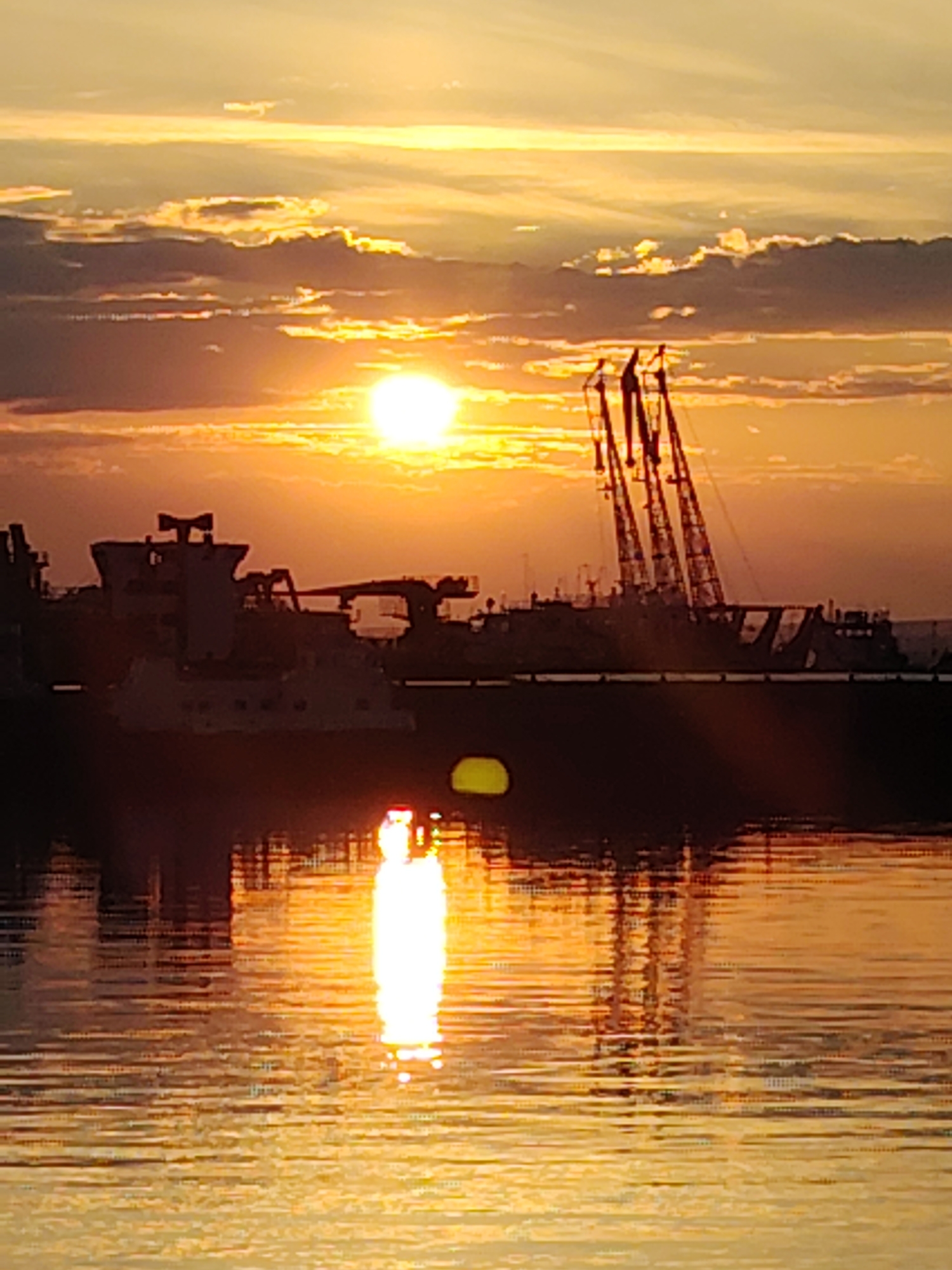 The image depicts a sunset over a body of water, likely the Kama River, near Chaikovsky, Russia. Silhouetted against the golden sky are industrial vessels, including a barge with multiple cranes. The setting sun casts a bright reflection on the rippling water, creating a shimmering effect. The clouds are illuminated by the sun, adding to the dramatic ambiance of the scene. The overall impression is one of industrial activity set against a beautiful natural backdrop.