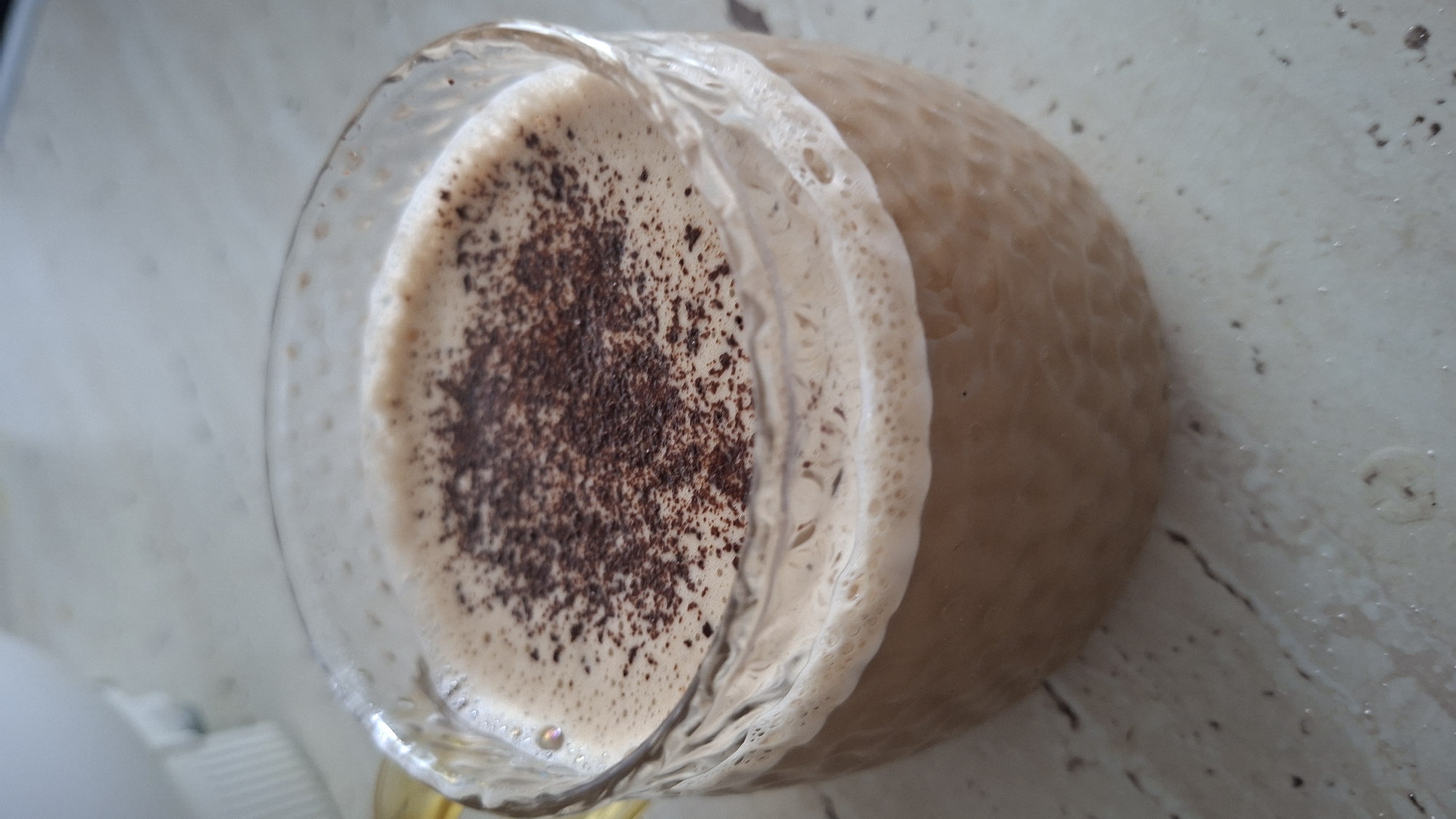 A close-up, overhead shot shows a clear glass mug filled with a frothy beverage. The mug is textured and appears to be on a speckled countertop. The beverage is a light brown color, topped with a layer of foam, and dusted with dark brown powder, possibly cocoa or coffee grounds. The background is blurred, but a white object with vertical lines is partially visible on the upper left. The scene depicts a beverage served in a mug, with no discernible location within the city of Myadzyel, Belarus, or any individuals present.