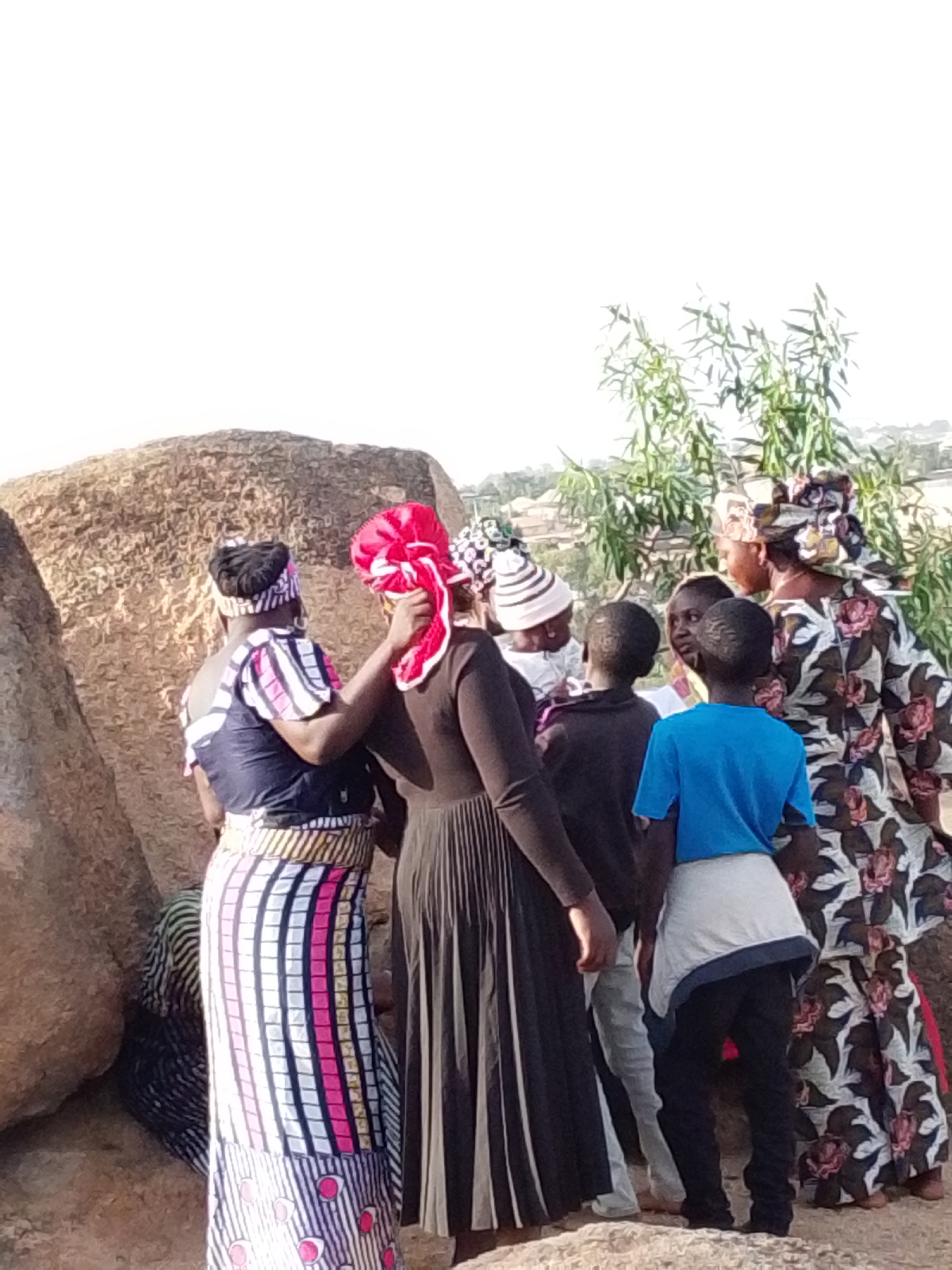 The image depicts a group of people, primarily women and children, gathered outdoors in what appears to be Barakin Akawo, Nigeria. The setting is characterized by large, rugged rock formations that occupy the left side and background of the scene. To the right, green foliage from bushes or trees is visible, along with hints of distant structures or buildings.
Several individuals are closely grouped together. In the foreground on the left, a woman is seen from behind, wearing a long, vertically striped skirt in shades of white, pink, black, and yellow, combined with a dark top and striped sleeves. Her hair is styled or covered with a dark wrap. Next to her, another woman, dressed in a dark gown, is either adjusting or having a bright red head covering adjusted on her head.
Further to the right, an adult woman in profile wears a distinctive patterned dress with floral designs in black, white, and pink, complemented by a matching headwrap. She appears to be engaging with other members of the group. Several children are present, including two boys seen from behind: one in a vibrant blue t-shirt and light shorts, and another in a dark top. Another child with a striped head covering is visible among the adults.
The group's arrangement suggests a social gathering, a communal interaction, or an observation of a shared event. The lighting indicates a clear, sunny day, likely late morning or afternoon, with soft shadows. The scene portrays a sense of community amidst a natural, rocky landscape. No text is visible in the image.