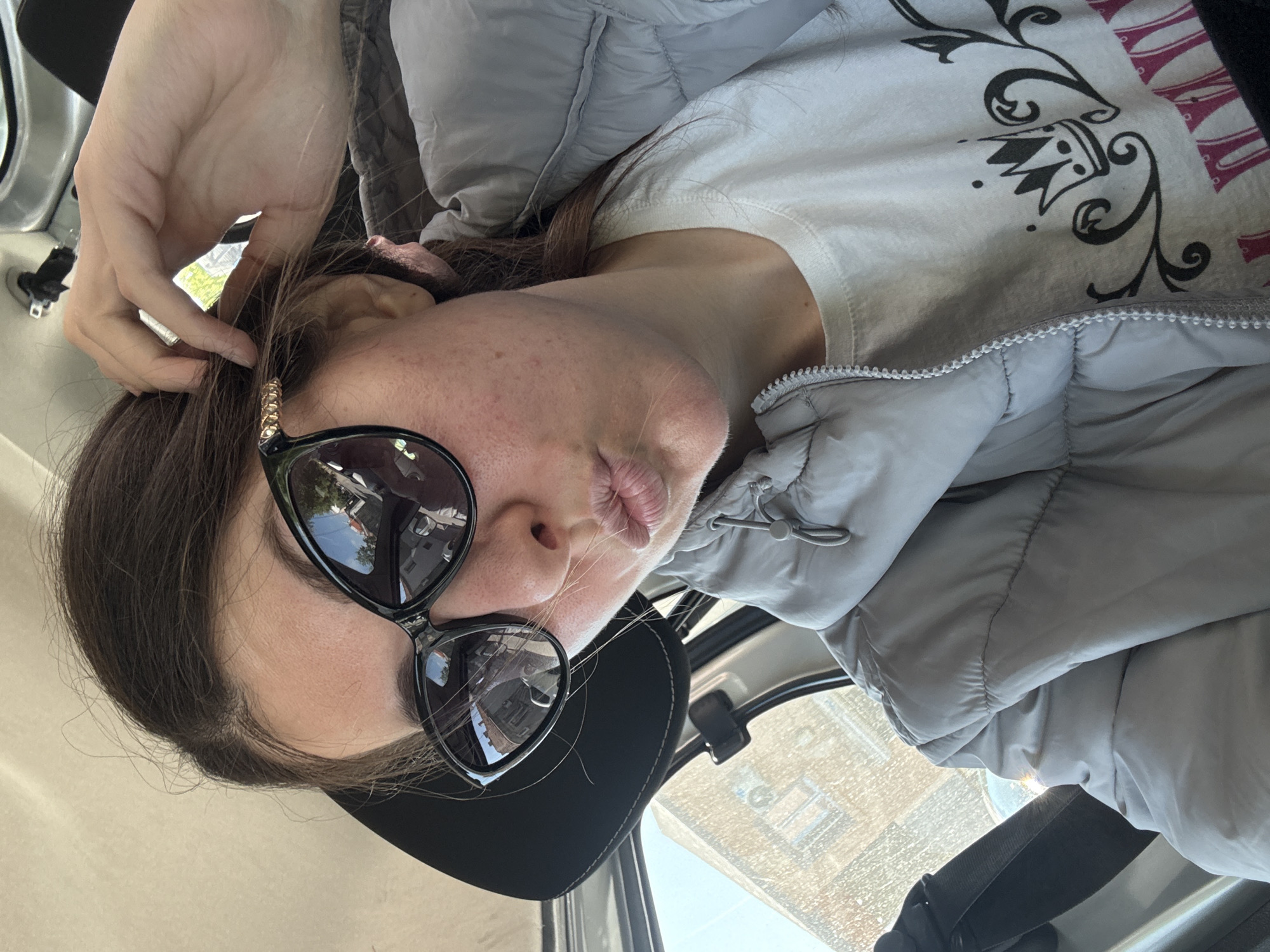 A woman is seated in a car, wearing a light gray puffer jacket, a white t-shirt with black and red designs, and black sunglasses with decorative gold accents. She is wearing a black baseball cap. Her lips are pursed, and her hand is near her hair. The reflection in her sunglasses suggests a street scene, including buildings. The car's interior is visible, including a window with raindrops on it. The city is unknown.