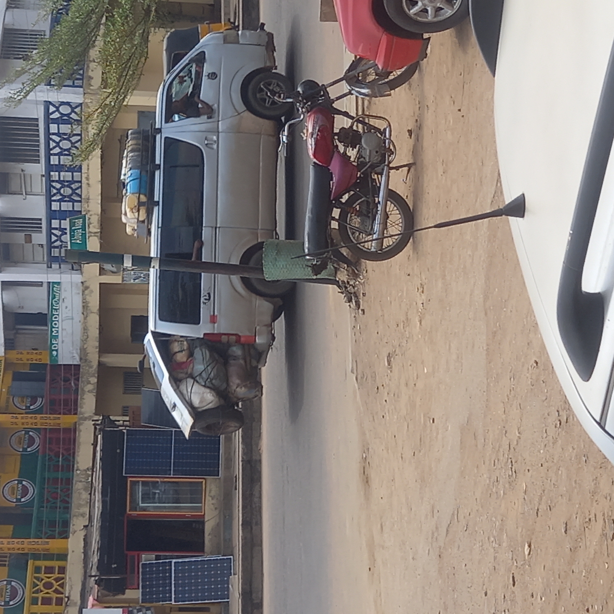 This street scene in Dadi Riba, Nigeria features a light-colored van heavily loaded on the roof and in the back. The van is parked on the side of the road next to a green signpost indicating "Abuja Road" and "DE MODELOutfit." Parked nearby are a red motorcycle and the front of a white car. The street surface is dirt. There are stacked crates and solar panels on the side of a building.