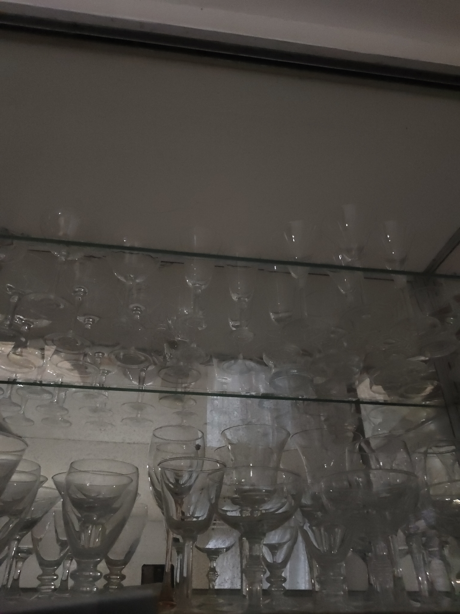 The image shows an indoor scene, possibly a cabinet or display shelf, filled with various glassware, primarily wine glasses and other stemmed goblets. The lighting is dim, and the objects appear to be made of clear glass, some with etched patterns. The shelves are made of glass. The scene is clean and orderly, with no people present. The location is identified as Davao City, Philippines.