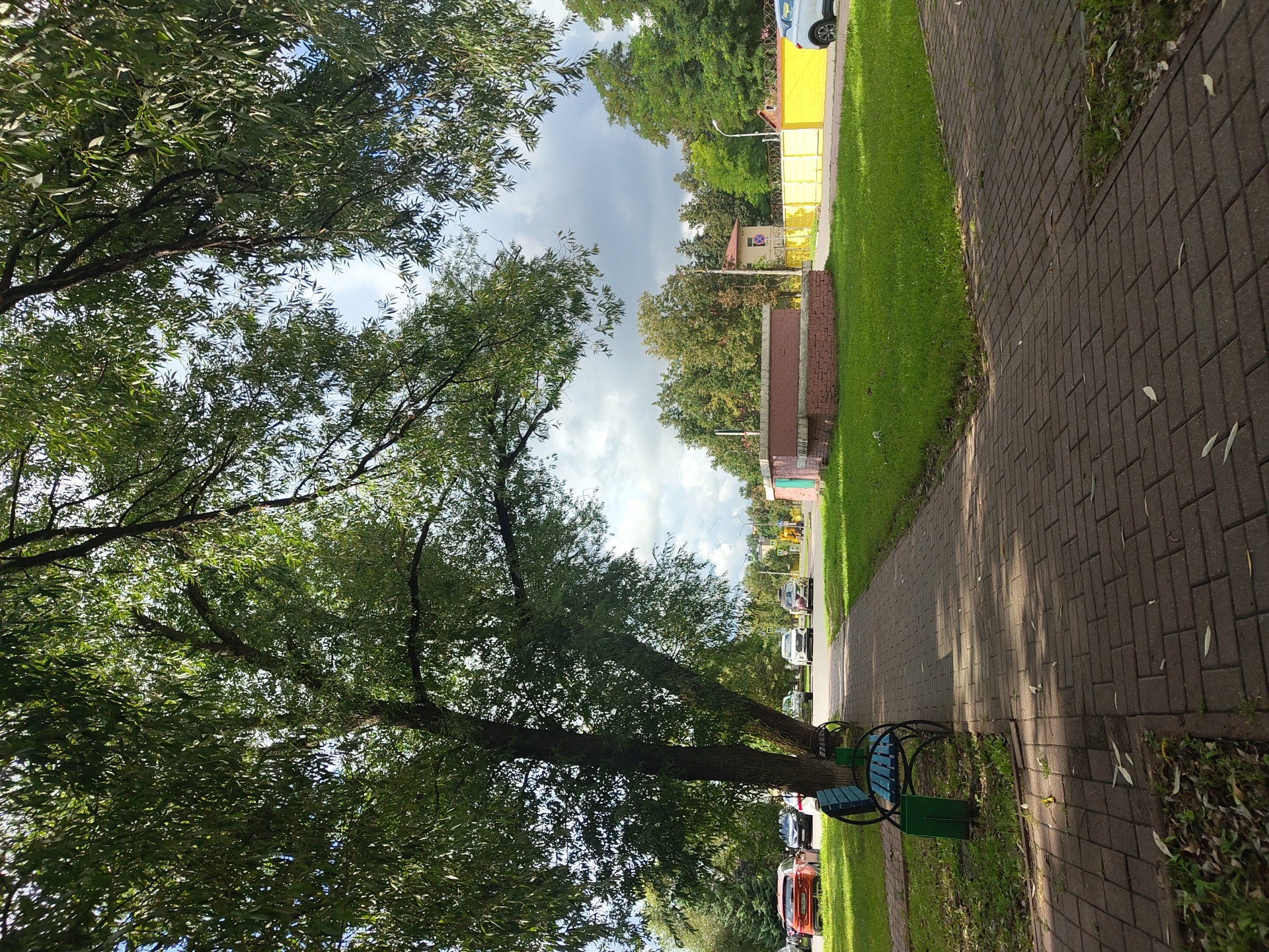 The image depicts a paved pathway in what appears to be a park or public space in Ramenskoye, Russia. The pathway is made of dark red brick pavers and is bordered by a grassy area. To the left of the pathway, a blue park bench with a black metal frame is situated. Further left, several cars are parked on a street. A large tree with dense green foliage extends into the upper portion of the image, casting shadows on the pathway. In the background, a low brick building with a pink facade is visible, adjacent to a bright yellow fence. Trees and a cloudy sky complete the scene.