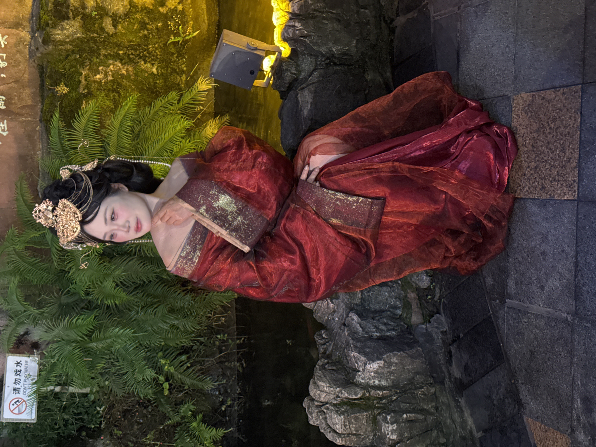 A woman dressed in a red, flowing garment with gold accents and an elaborate headdress sits on a rocky ledge. She is positioned to the right of the frame, looking to her left. Behind her, lush green ferns cascade downwards towards a dark body of water. A sign with Chinese characters and the English text "DON'T PLAY WATER" is visible on the left side of the image. The background features a textured wall illuminated by yellow light, with a spotlight visible on the right. The ground in the foreground is paved with dark stones. The overall scene appears to be an outdoor setting, possibly a park or garden in Yuzhong District, China, at night.