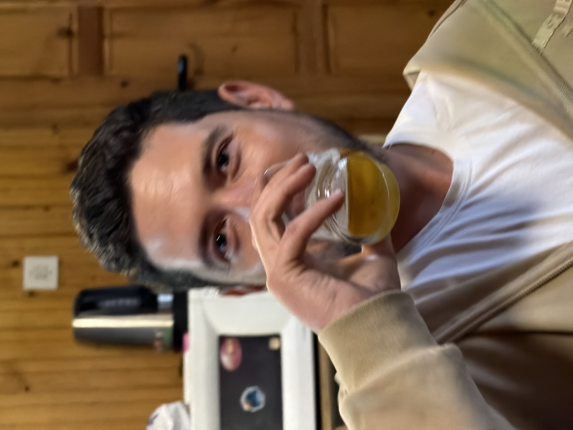 A man is drinking from a glass of yellow liquid. He has short dark hair, fair skin, and is wearing a white t-shirt and beige jacket. The background is a wood-paneled wall and a white object with a dark screen. The location is Lilkovo, Bulgaria.