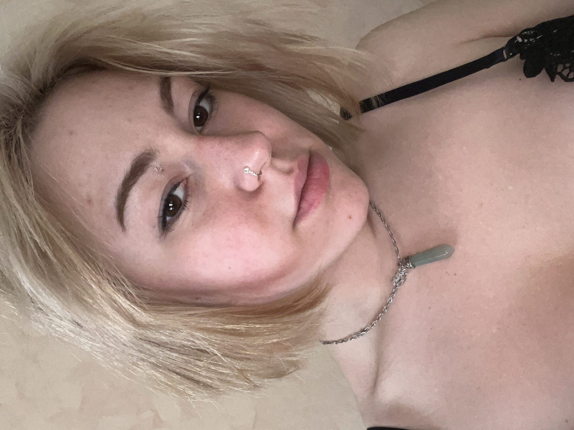 A close-up, angled portrait captures a young person, likely female, with short, light blonde hair framing her face. She has fair skin, with visible natural blemishes, and is looking directly at the viewer with a neutral, contemplative expression. Her dark brown eyes are prominent, enhanced by subtle eyeliner, and her eyebrows are well-defined. Notable facial features include a silver hoop piercing in her right nostril and a small, clear stud piercing positioned above her left eyebrow. Her lips are naturally pink and slightly parted.
The subject wears a black garment, appearing to be a bra or a camisole, with a delicate black lace detail visible on the strap over her right shoulder. Around her neck, a thin silver chain necklace is worn, adorned with a light green, pointed pendant, possibly made of stone or crystal, which appears to be wrapped with silver wire at its top. The background is a plain, light, possibly beige-colored surface, intentionally blurred to keep the focus on the subject. The image's angle suggests the person is reclining or lying down while posing for the photograph. No specific activity is depicted other than posing. This scene is situated in Odesa, Ukraine.