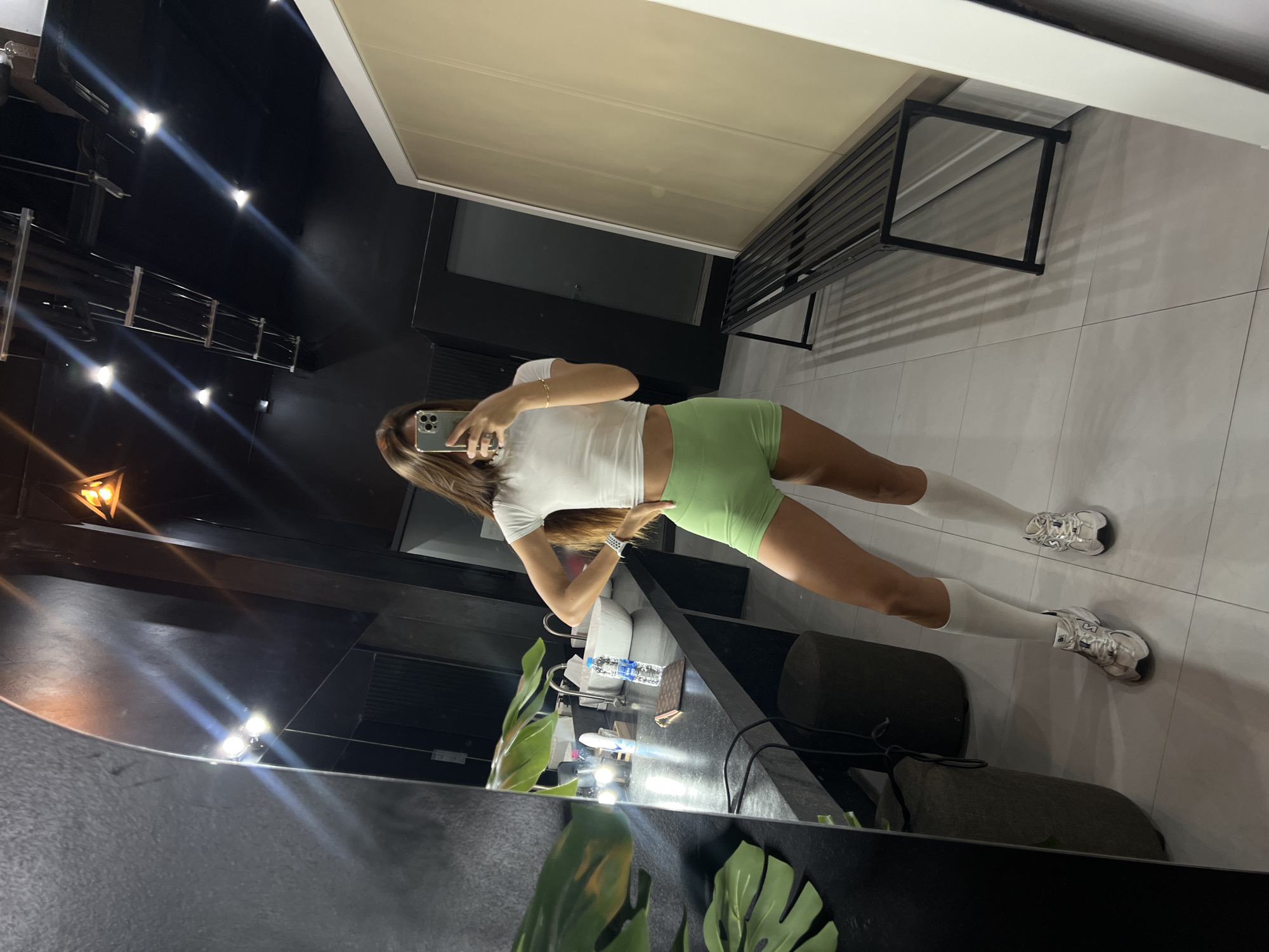 In a dimly lit, modern changing room, a young woman with long brown hair stands in front of a large mirror, taking a selfie with her smartphone. She is wearing a white cropped t-shirt, light green athletic shorts, white knee-high socks, and silver athletic shoes. Her left hand rests on her hip, and her right hand holds the phone. The room features a dark countertop with sinks and a water bottle, as well as a long black metal bench. The floor is tiled, and the walls are dark, with some lighting fixtures visible on the ceiling.