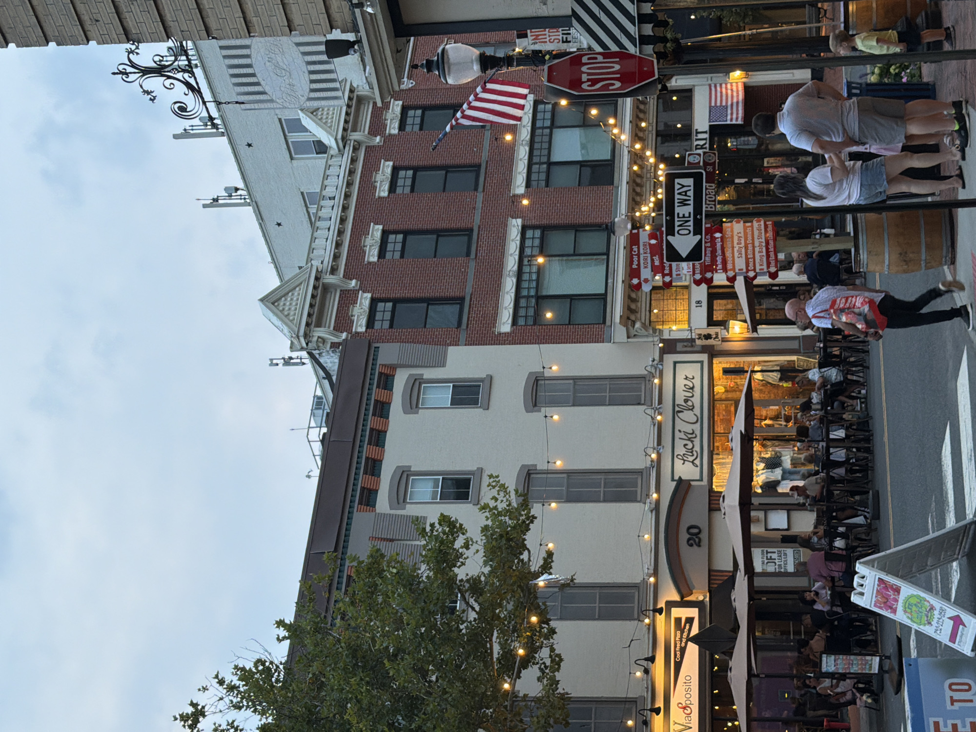 The image captures a street scene in Red Bank, New Jersey, United States of America, during the late afternoon or early evening. The street is lined with two- and three-story buildings, showcasing a mix of architectural styles. The building on the left is a lighter cream color with arched windows on the upper floors and a row of storefronts at street level, one of which is labeled "Lucky Clover." String lights are strung across the street between the buildings, illuminating the scene with warm, yellow light.

On the sidewalk, tables and umbrellas are set up for outdoor dining in front of some of the establishments. Several people are seated at these tables, engaged in conversation or eating. Pedestrians are visible on the sidewalk and crossing the street. A man in a white shirt and red striped bag is walking towards the viewer, while a family of four stands on a corner near a street lamp and a "STOP" sign. A young boy in a yellow shirt is looking to the right.

Various signs are posted along the street. One sign indicates "ONE WAY" with an arrow pointing to the left, and a cluster of smaller signs list various businesses and street names, including "Broad St." and "18." An American flag is displayed on the facade of the brick building on the right. The sky above is overcast with soft clouds. The overall atmosphere is one of a bustling urban environment with a charming, pedestrian-friendly street.