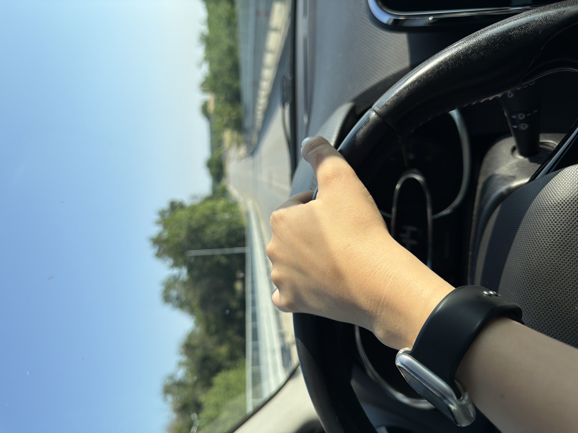 The image captures a first-person perspective from inside a car, looking forward and slightly to the left. The primary subject is a person's right hand and forearm, gripping the top-right portion of a black steering wheel. A smartwatch with a black band and a silver-colored face/clasp is worn on the wrist.
Through the car's windshield, a clear blue sky dominates the upper-left part of the frame, indicating a bright, sunny day. Below the sky, a road is visible, stretching into the distance with white lane markings. Lush green trees line the right side of the road, suggesting a natural or semi-natural environment, possibly a highway or rural road. The background outside the car appears slightly blurred, emphasizing the foreground elements.
Inside the car, parts of the dashboard and instrument cluster are faintly visible behind the steering wheel. The steering wheel itself appears to have some texture or wear on its surface. The overall impression is of someone driving, with the hand firmly on the wheel. The image itself is rotated 90 degrees clockwise, making the horizon appear vertical.