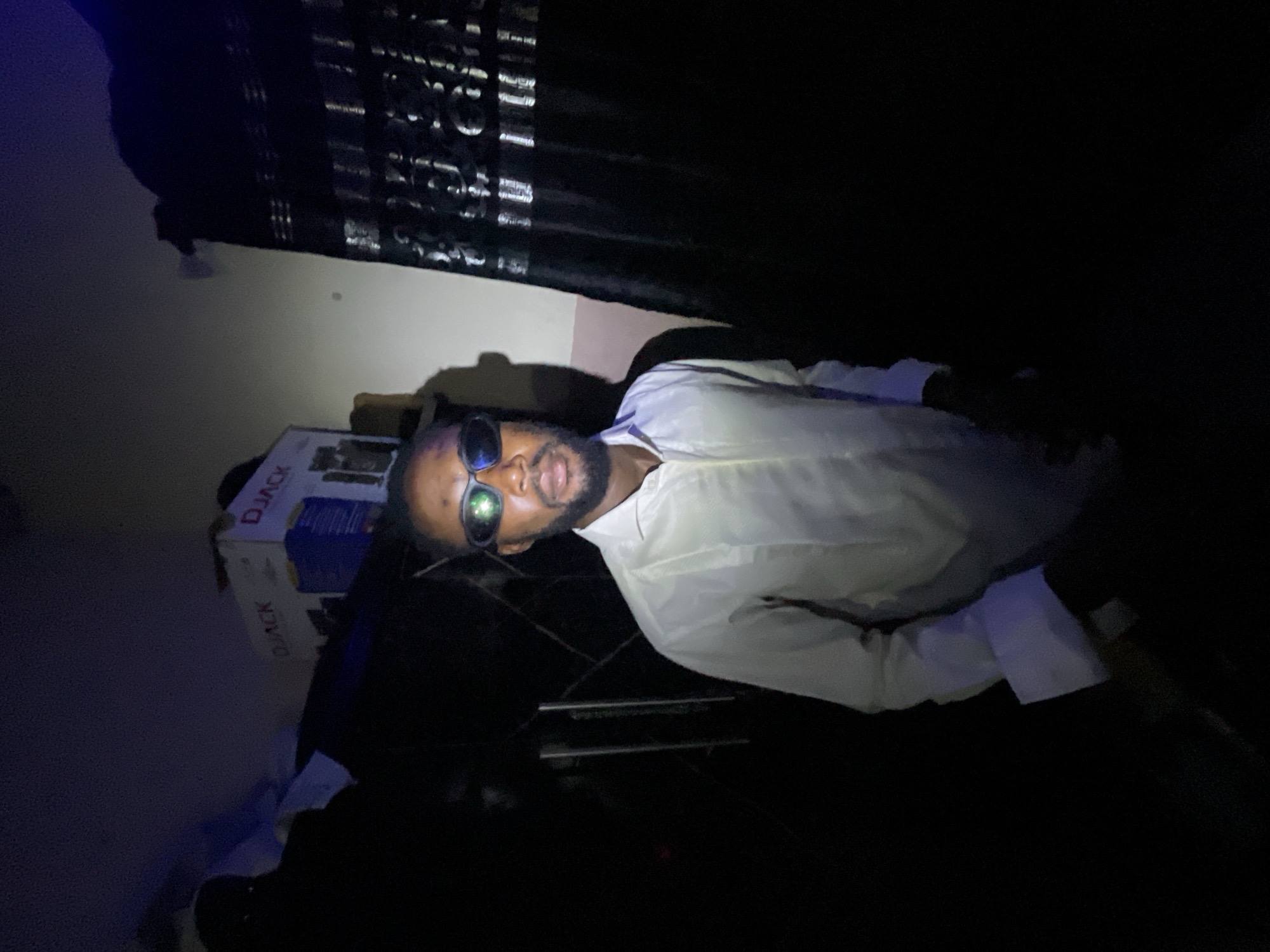 The image appears to be taken indoors, possibly in a private residence in Lokoja, Nigeria. The lighting is poor, with much of the scene obscured by darkness. A Black man is visible, wearing sunglasses and a light-colored shirt. The man has a beard. Behind him is a box with "DJACK" written on it. The background includes what looks like a curtain. There is no specific information about the location in Lokoja, Nigeria to be seen.