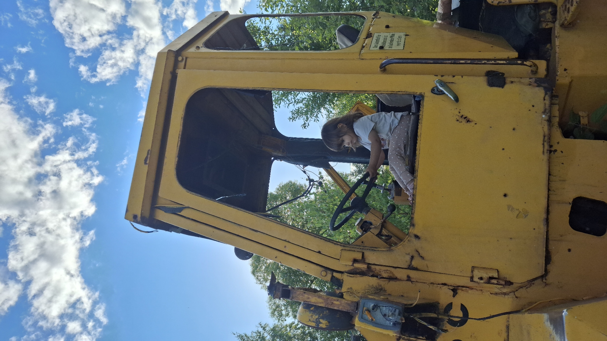 A young girl sits in the driver's seat of an old, yellow, industrial vehicle, likely a loader or similar construction equipment, with her hands on the steering wheel. She is wearing a light-colored t-shirt and patterned leggings. The vehicle's interior is visible, showing the steering wheel, a seat, and some controls. The exterior of the vehicle is weathered and shows signs of rust and age. The scene appears to be outdoors, with green trees and a bright blue sky with scattered white clouds in the background. There is a sign with German text on the side of the vehicle. The image is captured in a low-angle, vertical orientation, with the sky occupying the upper portion of the frame. This image was taken in Innolovo, Russia.