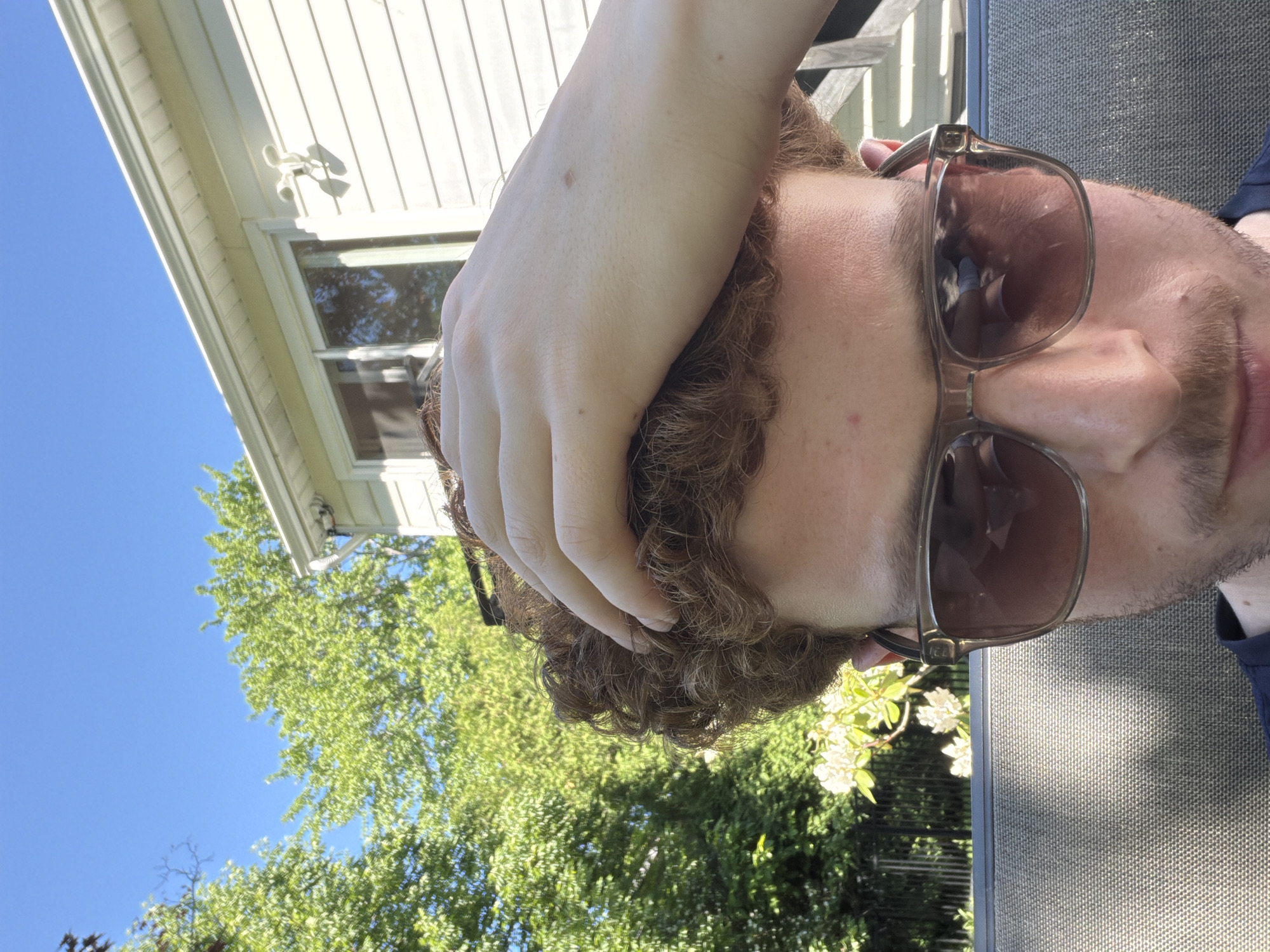 The image is an outdoor close-up, rotated 90 degrees clockwise, showing a person against a bright, sunny background in Pepper Pike, United States of America.
**Subjects:** The primary subject is a person, likely male, with fair skin, curly reddish-brown hair, and light brown or tan-framed sunglasses. Their right hand rests on top of their head, fingers gently spread, partially obscuring their hair. The sunglasses cover their eyes, reflecting bright outdoor light and what appears to be a light-colored object or clothing. Part of their ear and the side of their face are visible, along with a light beard or stubble around their chin and upper lip.
**Scene and Setting:** The scene is outdoors, likely a residential backyard or patio. In the upper left, a portion of a white-sided house with a window is visible. A small white light fixture or security camera is mounted on the siding above the window. A clear, bright blue sky fills the upper left and central background. Lush green trees and foliage are prominent, especially in the lower half of the image, suggesting a verdant environment. Some clusters of small white flowers are visible in the lower right, near the person's shoulder. To the right of the person, a textured, dark gray surface, possibly part of a speaker or outdoor furniture, is visible.
**Activity or Event:** The person appears to be relaxing outdoors, possibly enjoying the sunshine. Their hand on their head might indicate a casual pose, a moment of thought, or simply resting.
**Notable Details:** The bright sunlight, clear blue sky, and distinct shadows (e.g., on the house and reflected in the sunglasses) indicate it is daytime, likely mid-morning or afternoon. The weather is clear and sunny. No visible text is present in the image. The overall mood appears calm and relaxed.