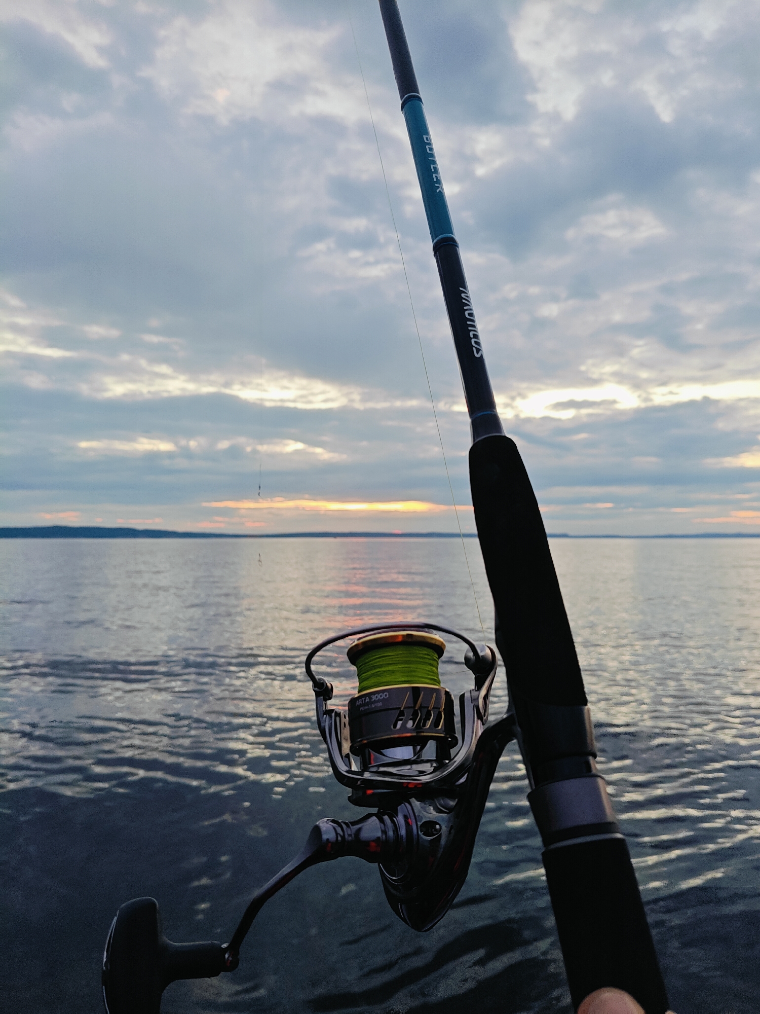 This media file features a close-up of a fishing rod and reel positioned over a body of water, likely a lake or calm sea, under a cloudy sky at dusk. The foreground is dominated by the fishing gear, with the dark handle of the rod and the metallic reel clearly visible. The reel has a spool of bright green fishing line. The rod itself has text visible: "BUTLER" on a turquoise section and "MAVICROS" on a darker section.
In the background, the water stretches to the horizon, where a soft, diffused light suggests the setting sun, casting a warm glow on the clouds and the water's surface. The sky is filled with dramatic, layered clouds, some tinged with orange and pink from the sunset, while others remain a soft grey. A single fishing line extends from the rod into the water, hinting at the activity of fishing. The overall mood is serene and tranquil, evoking a sense of peacefulness associated with sunset fishing. The location is identified as Chaikovsky, Russia.