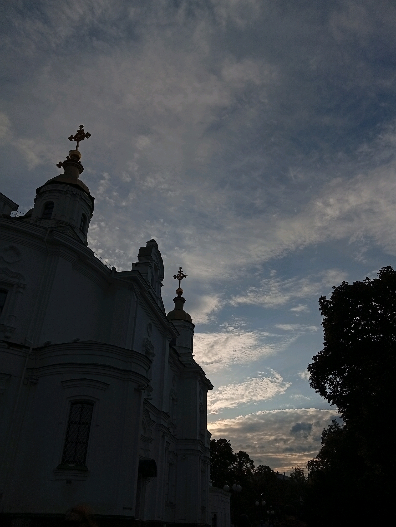 The image shows a low-angle shot of the facade of a white building with golden crosses on its domes, possibly a church or monastery, against a cloudy sky. The building is silhouetted in the foreground, with details of its architecture, including windows and decorative elements, visible despite the low light. The sky is filled with various cloud formations, ranging from wispy streaks to more dense formations, with hints of sunlight breaking through. A large, dark tree is visible on the right side of the frame, further contributing to the silhouette effect. The scene appears to be captured at dusk or dawn due to the lighting conditions and the dramatic sky.