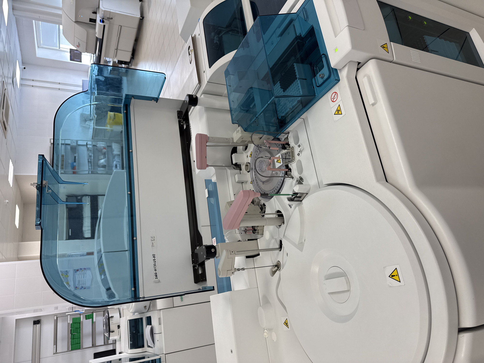 The image captures a clean, well-lit indoor laboratory setting, showcasing advanced automated laboratory equipment. The primary focus is on a large, white analytical machine featuring transparent blue protective covers. Visible through the covers are intricate internal components, including what appears to be a sample carousel for vials and robotic probes, suggesting it's an automated analyzer for chemical or biological samples.
The laboratory walls are tiled white, and overhead fluorescent lighting illuminates the space. Other large, white laboratory instruments are visible in the background, implying a comprehensive testing facility. A window in the upper left background indicates natural light may also be available.
Small green indicator lights on the equipment suggest that the machines are powered on and operational. Several warning labels are present on the machinery: yellow triangular stickers with black exclamation marks (general warning), and a circular sticker with a red line through it alongside a biohazard symbol (indicating biohazard risk and a prohibition). A white label with the text "2086-01 (32-05-01)" is affixed to the side of one of the blue covers. No people are visible, highlighting the automated nature of the processes. The overall impression is one of a modern and sterile clinical or research laboratory, located in Moscow, Russia.