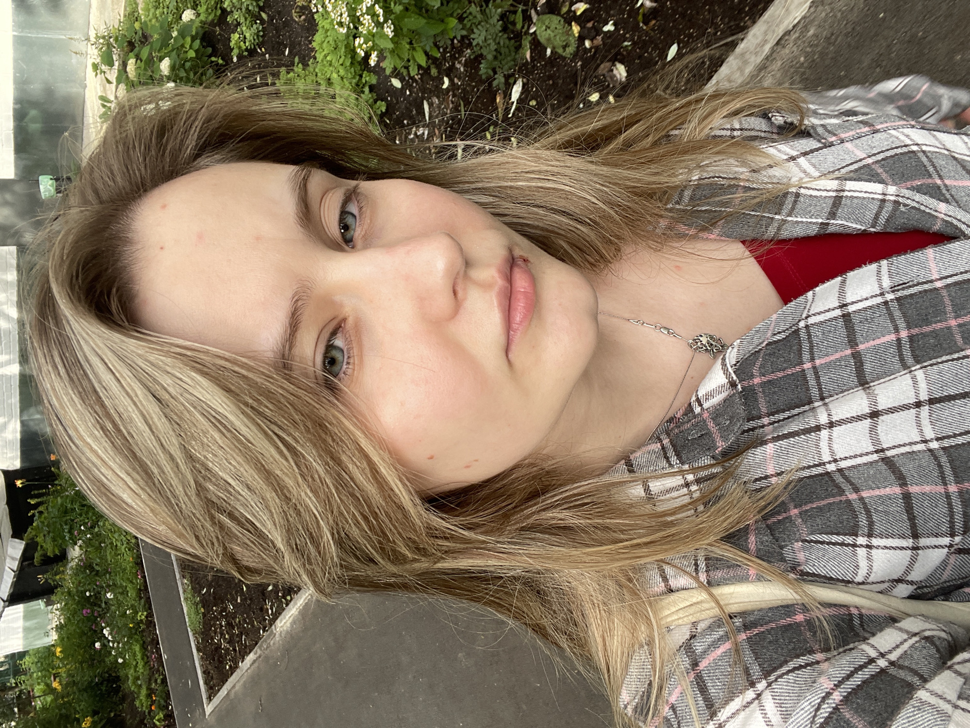A young woman with blonde hair and blue eyes is taking a selfie outdoors. She is wearing a plaid shirt over a red top and a silver necklace. The background shows a garden with green plants and flowers, and a paved walkway. The woman's expression is neutral, and she is looking directly at the camera.