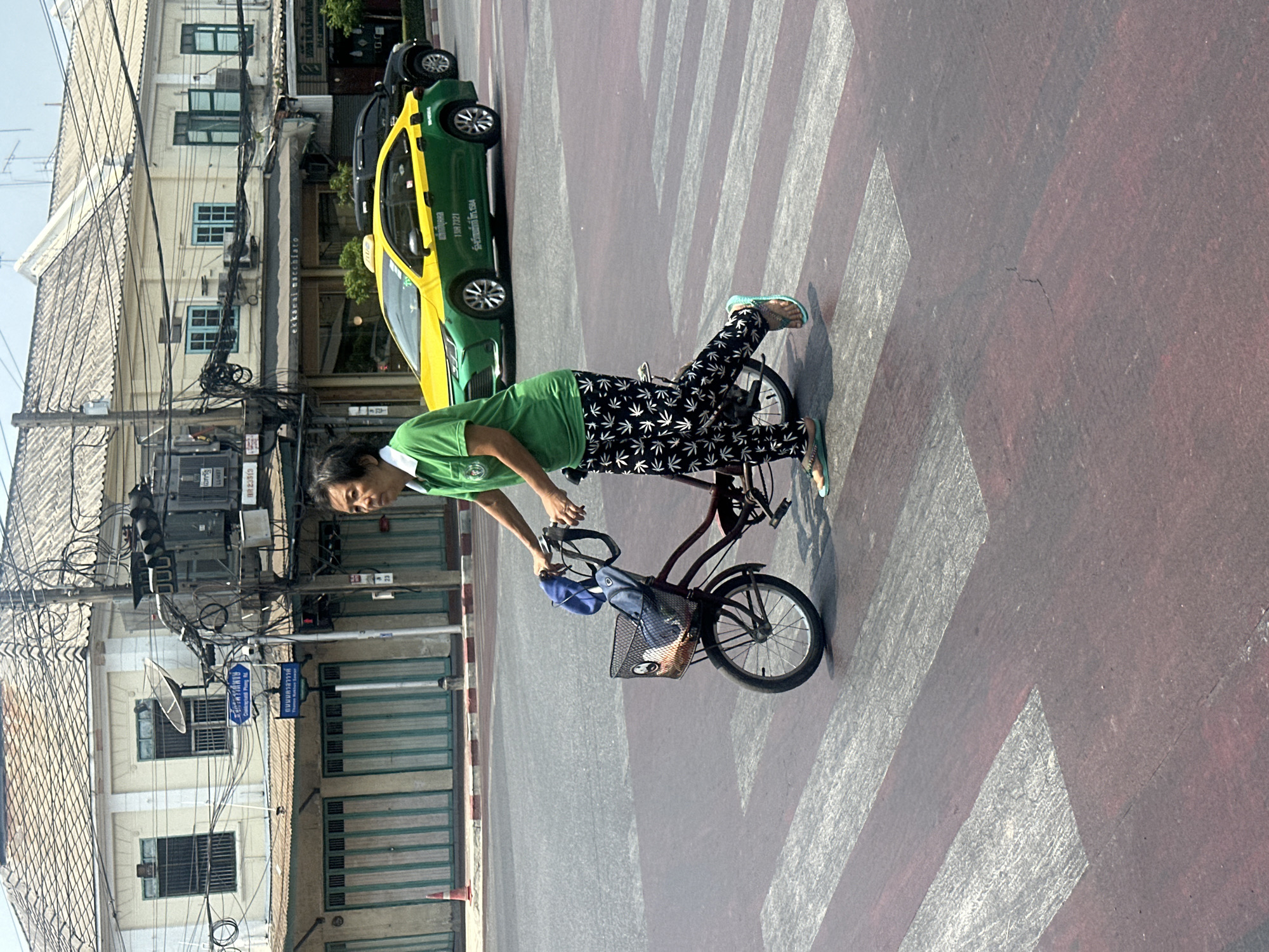 A pedestrian is crossing a Bangkok, Thailand, street riding a small bicycle in an area marked with alternating red and white crosswalk stripes. The pedestrian is wearing a green shirt and black pants with white marijuana leaves printed on them. The bicycle has a small basket on the handlebars. In the background are city buildings, street lights, and a green and yellow taxi. Street signs are visible in Thai script. The day is bright and sunny.