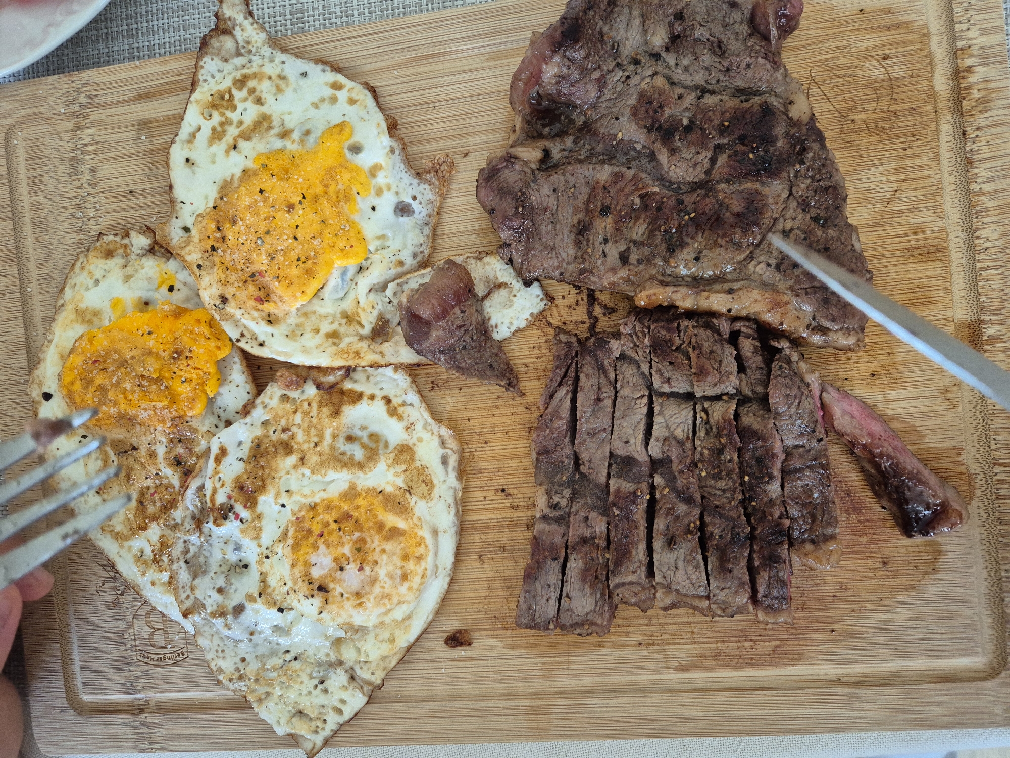 A top-down, eye-level, medium shot captures a wooden cutting board laden with a meal. The board is positioned on a neutral-toned woven placemat. In the upper portion of the frame, three fried eggs are arranged, their yolks varying in consistency from fully cooked to runny, and all lightly seasoned with salt and pepper. A silver fork, held by an unseen individual, is poised above the eggs, suggesting the meal is about to be eaten. Below the eggs, a sliced steak occupies the lower half of the board. The steak is cooked to a medium-rare, with visible pink centers in the slices. Its surface is seasoned with pepper and possibly other spices. A silver knife rests at the bottom right of the board, its tip angled towards the steak. The lighting appears to be natural, casting soft shadows. The meal is served on a bamboo cutting board. There is no discernible information or context within the image to indicate a specific location in the city of Varna, Bulgaria.