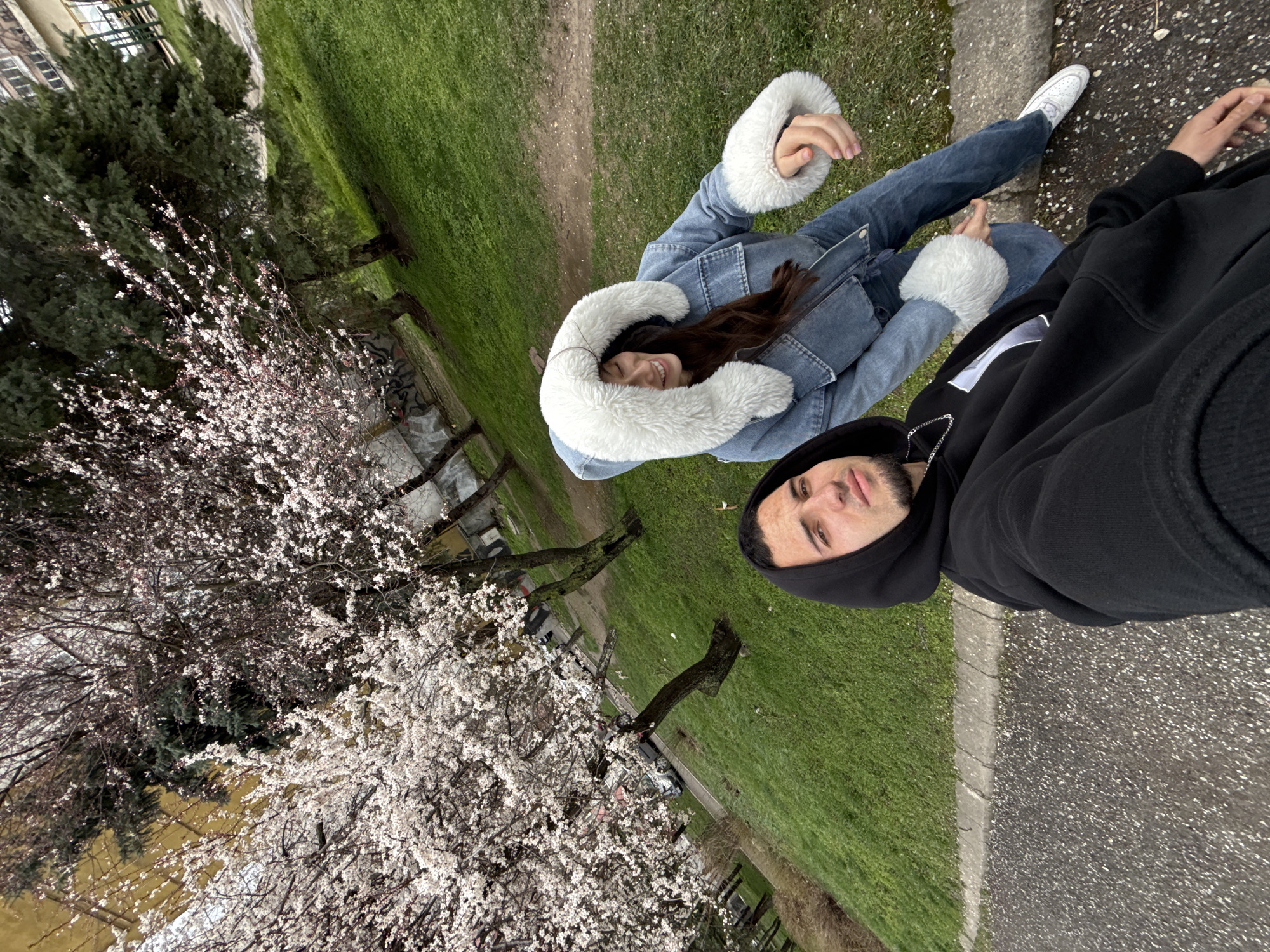A man and a woman are in an outdoor setting near the city center in Shumen, Bulgaria, as indicated by background elements. The woman is wearing a denim jacket with white fur trim. The man is wearing a black hoodie. They are standing near a blooming tree, likely in a park or green space. The ground appears to be covered in grass with a paved walkway visible. The perspective suggests the image was taken as a selfie.