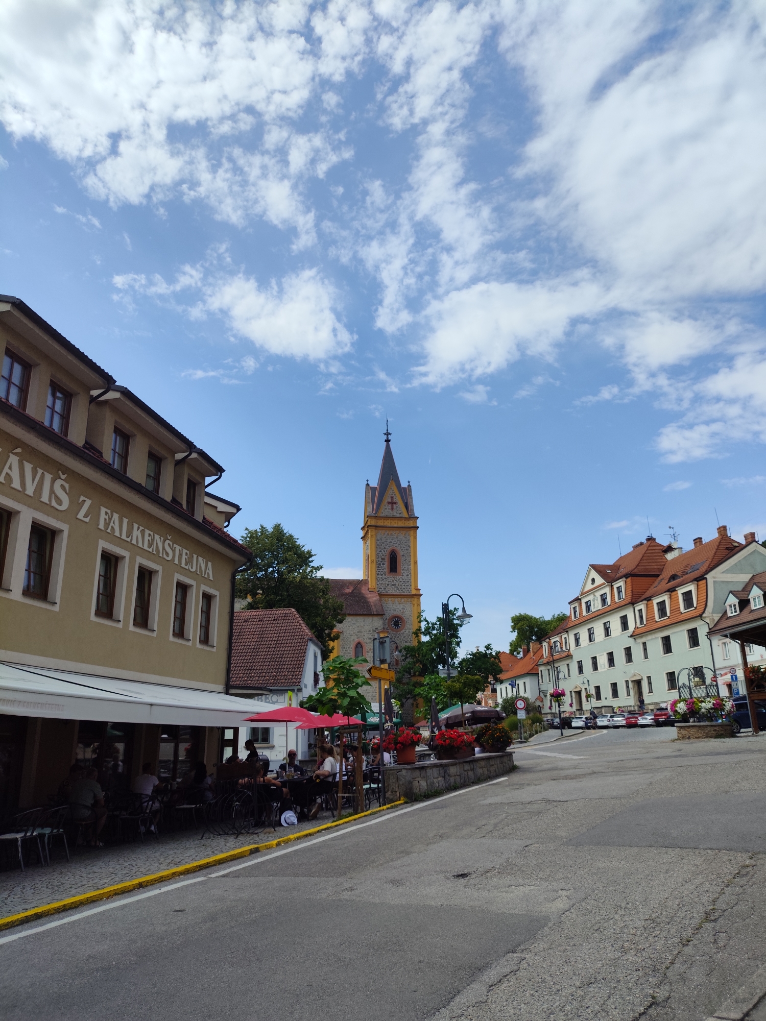 The image depicts a street scene in Hluboká nad Vltavou, Czechia, featuring a row of buildings lining a curved street. On the left, a light yellow building with a white awning displays signage with text that appears to be "VÁVIŠ Z FALKENŠTEJNA." Outdoor seating with umbrellas and tables is set up along the sidewalk in front of this building, with several patrons seated and conversing. Further down the street, a prominent church tower with a red roof and a cross on top stands out against a blue sky with scattered white clouds. The street itself is paved and appears to be moderately trafficked, with parked cars visible in the distance. Other buildings with red-tiled roofs and light-colored facades are visible along the right side of the street, some adorned with flower boxes. The overall impression is that of a pleasant, historic town square or street on a clear day.