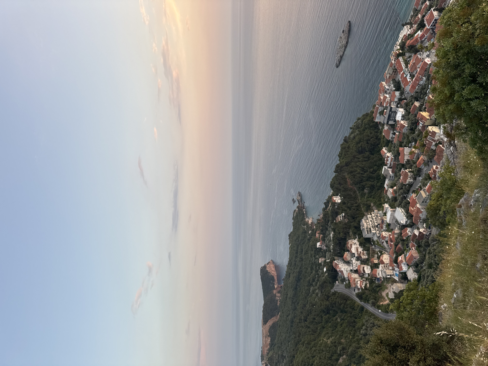 The image shows a coastal view in Sveti Stefan, Montenegro. The sky transitions from light blue to a mix of pale pink and orange near the horizon. The sea occupies the central portion of the view, its surface relatively calm with visible reflections.
A rocky islet is situated offshore. The coastline is characterized by dense green vegetation and clusters of buildings with red-tiled roofs. A road winds through the landscape. The perspective is from an elevated position. No people are visible.