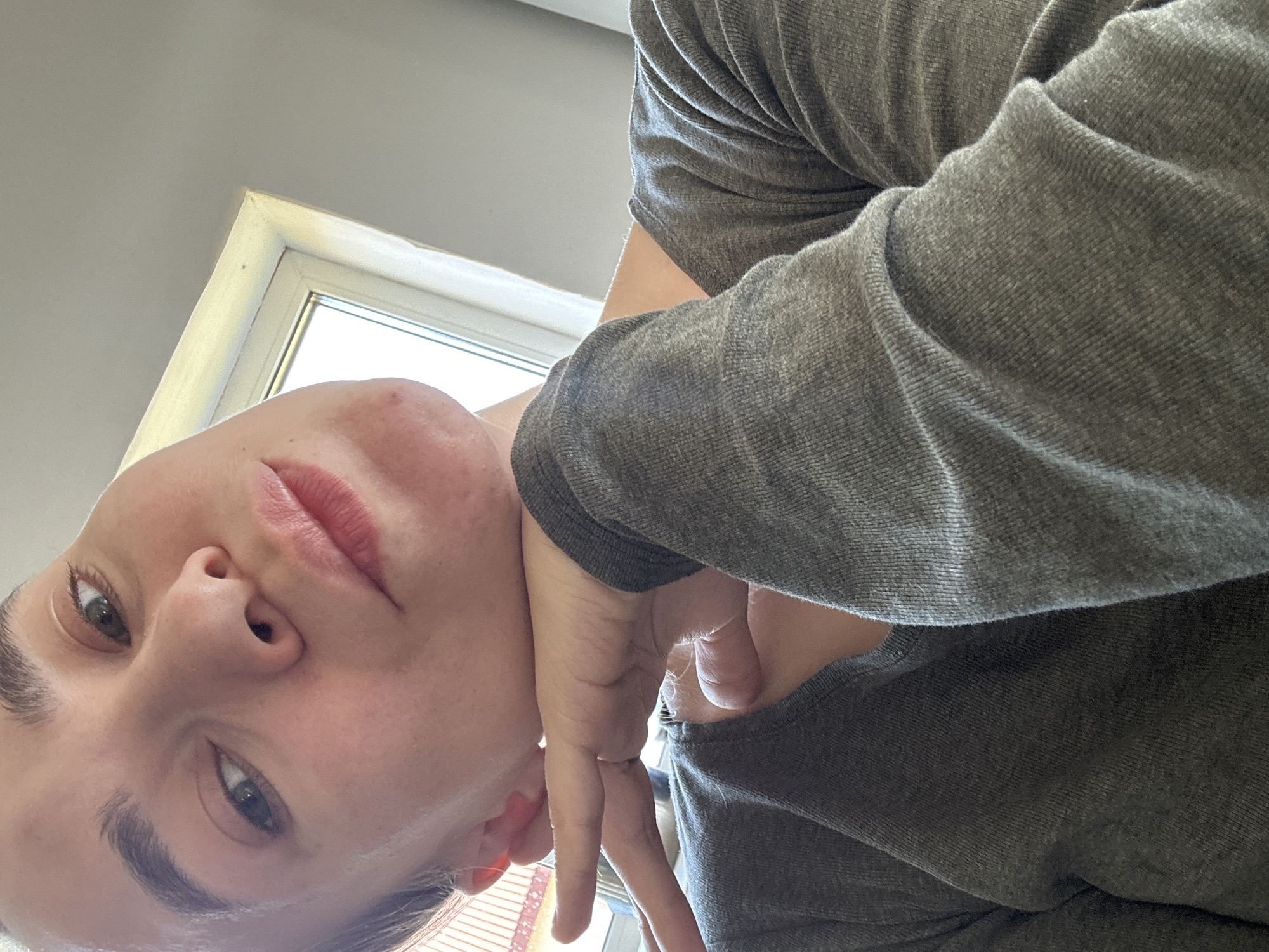 A selfie of a person with fair skin wearing a gray long-sleeved shirt. The person's head is tilted, positioning their face in the lower left corner of the frame. The background features a window frame and a light gray wall. A hand, also belonging to the person, is positioned near their neck, making a 'peace' sign with fingers. No information to determine the location in the city of Kharnavy, Russia is available.