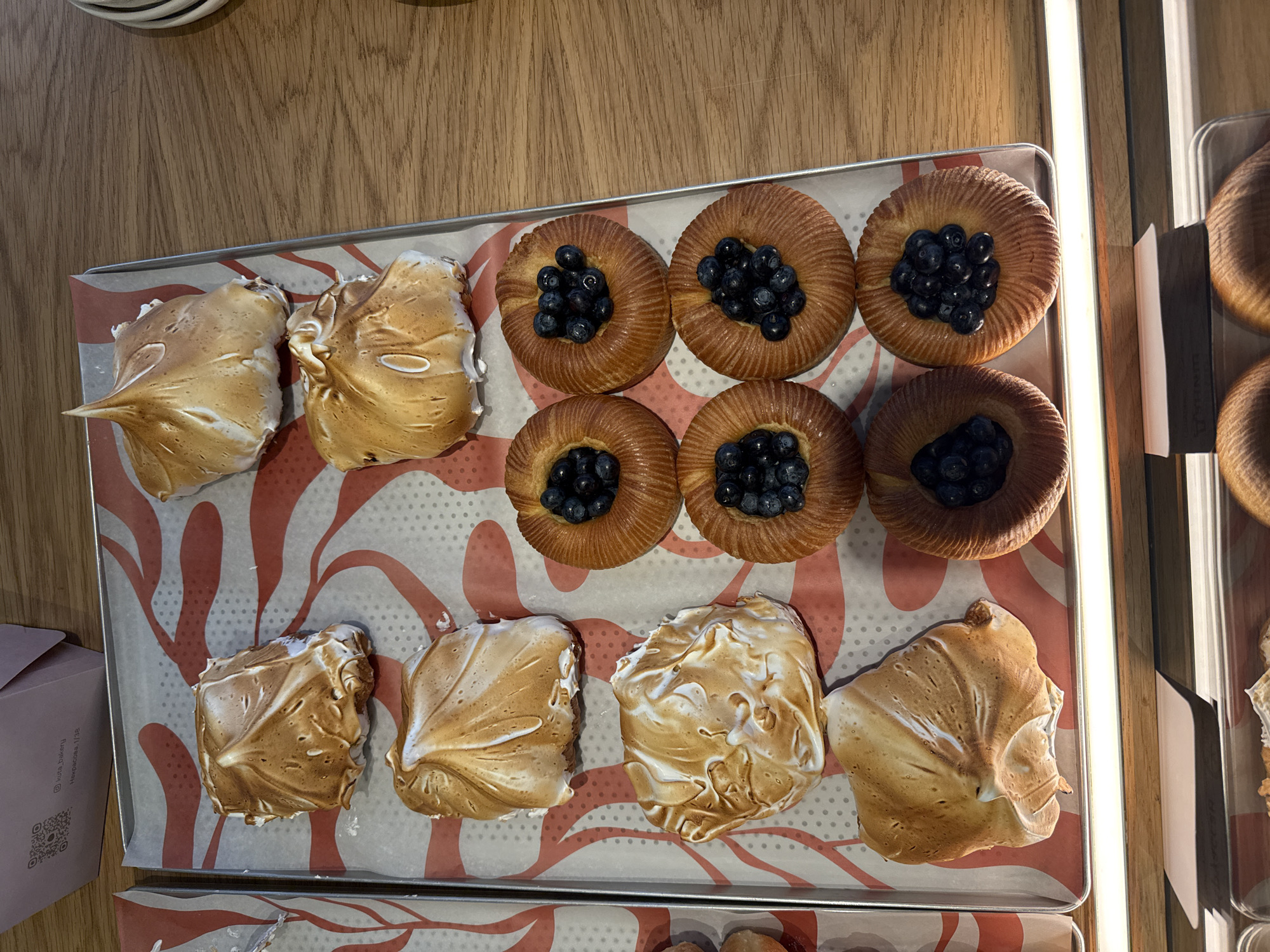 An overhead shot shows a metal baking tray filled with pastries on a wooden surface. The tray is lined with paper featuring a red wavy pattern. On the left side of the tray are four baked goods topped with meringue that has been browned. To the right of these are four round pastries, each filled with blueberries. The pastries are arranged in two columns and two rows. The background shows a light pink box with black text and a QR code. The scene appears to be an indoor setting, possibly a bakery. The location is Saint Petersburg, Russia.