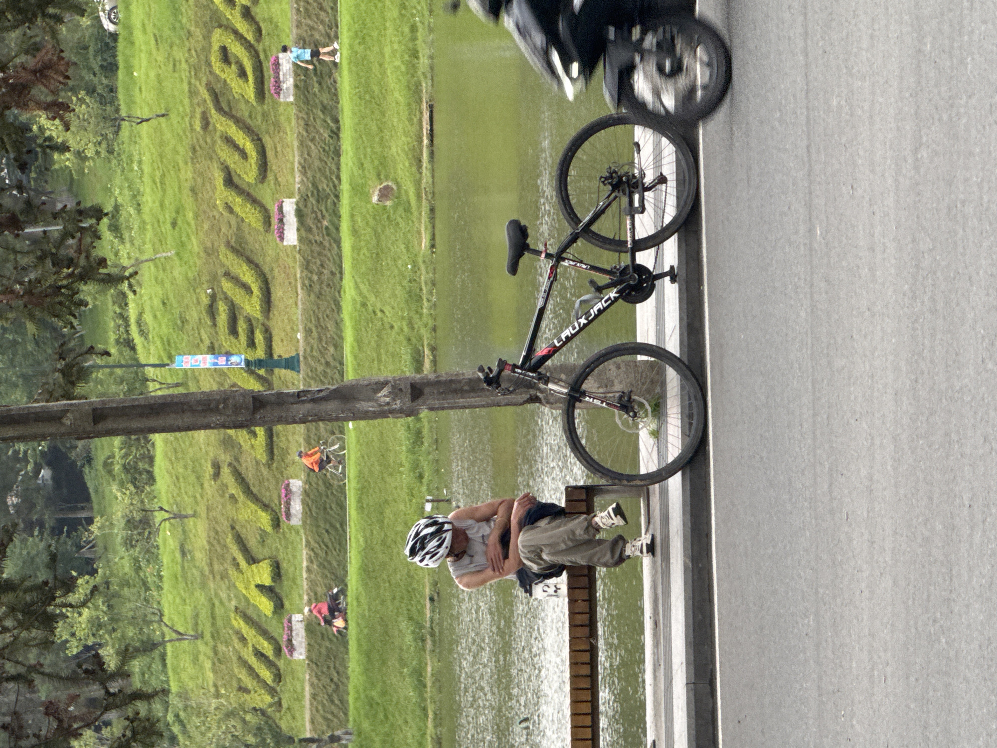 The image depicts a waterside scene in Da Lat, Vietnam, with a road in the foreground and a park across a lake. In the immediate foreground, a gray asphalt road is separated by a curb from a gray paved area. A black LAUXJACK mountain bike with red accents is parked upright on the pavement, leaning against a dark, rough tree trunk on the right. A blurred black motorcycle is partially visible, moving rightward on the road. To the left of the tree trunk, a person, wearing a white cycling helmet, a gray sleeveless shirt, and light-colored trousers with white sneakers, sits on a wooden slatted bench. The person's head is lowered, and a white cup or bottle rests on the bench next to them.
Across a body of green water, likely a lake, a landscaped green bank leads to a paved pathway. On this path, from left to right, a person on a red scooter, a person cycling in an orange top, and a person walking in a light blue shirt are visible. Above the path, a manicured green hillside features large, sculpted hedge letters spelling "KÝ YÊU TƯ ĐÀ," with preceding letters obscured on the left. Two rectangular planters with pink flowers are positioned along the path below the hedge letters. A small brown dog is visible on the green slope between the cyclists and the walker. Scattered green trees populate the hillside and the distant background. A lamppost with a blue banner is visible on the left side of the path.