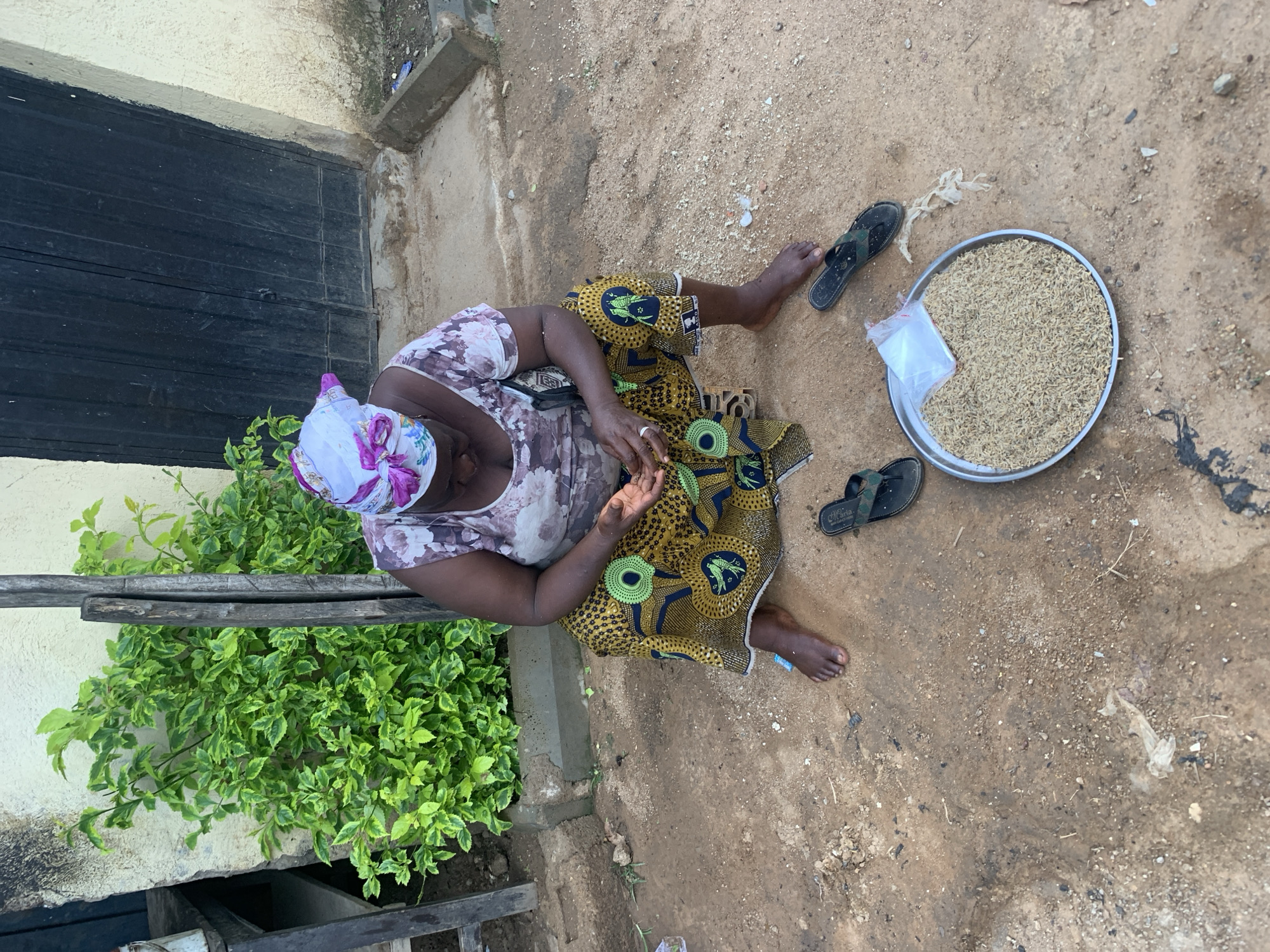 An adult woman sits on the ground in Jos, Nigeria. She wears a patterned dress, floral headwrap, and a ring on her left hand. She is sorting grains, possibly rice, in a silver tray. Beside the tray are two black sandals and a small plastic bag. She is seated next to a wall with a black wooden door and a potted plant. The ground is dirt and gravel.