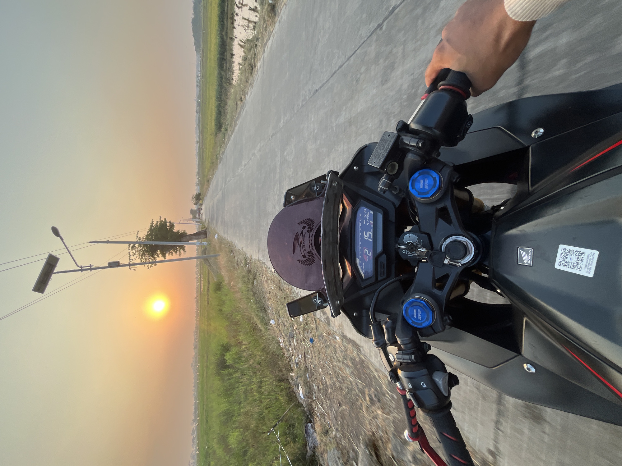 The image depicts a first-person perspective of someone riding a black motorcycle on a paved road in Pecangaan, Indonesia, during sunset. The road is flanked by lush green rice fields. A street lamp with a solar panel is visible on the left side of the road, along with a single tree. The motorcycle's dashboard displays a digital readout of "2 15" and "41449". The rider's hand is visible on the right handlebar, and their arm is clad in a light-colored sleeve. The sun is low in the sky, casting a warm glow over the landscape.