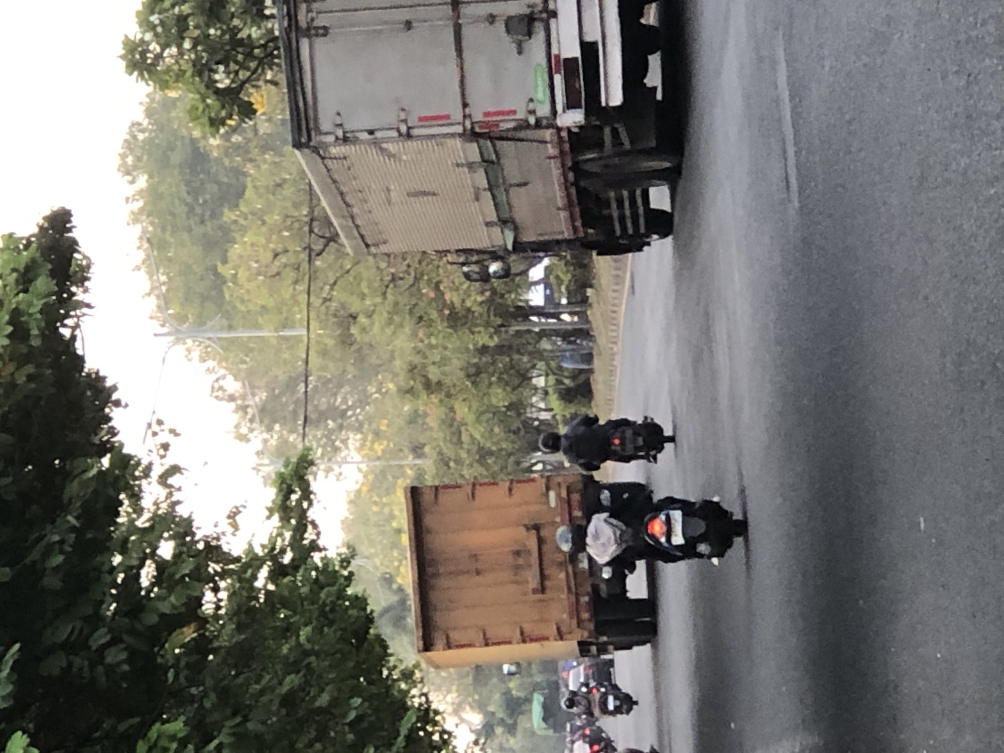 A busy street scene in Sumorame, Indonesia, is captured in this image. The foreground is dominated by a gray asphalt road with white lane markings. Several motorcycles are in motion, with two riders clearly visible in the center. One rider in front is wearing a light-colored hooded garment and riding a black motorcycle, while another rider behind them, wearing a dark jacket, is on a black motorcycle. To the left, more motorcycles are visible in traffic.
In the mid-ground, two large trucks are prominent. A large, weathered orange truck with its rear facing the camera is on the left side of the road. To its right, a silver box truck is also on the road, its side and rear clearly visible. Both trucks appear to be stationary or moving slowly.
The background is filled with lush green trees and foliage, suggesting a natural environment bordering the urban setting. The sky is bright and appears to be either early morning or late afternoon, with some atmospheric haze. The overall impression is a typical street view of Indonesian traffic, characterized by motorcycles and larger vehicles sharing the road amidst abundant greenery.