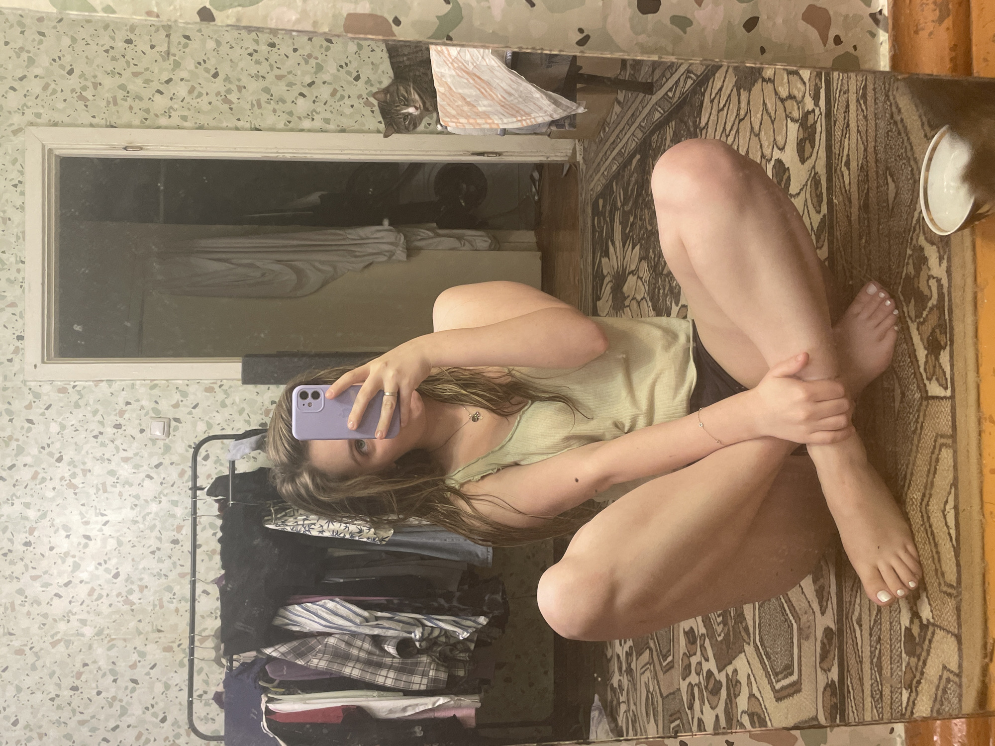 A young woman with long, light brown hair is sitting cross-legged on a patterned rug in front of a mirror. She is wearing a light green tank top and dark bottoms. She is holding a purple phone in her right hand, taking a selfie. Her left arm is resting on her knee, and her right arm is bent, with her hand near her face. A delicate bracelet with small charms is visible on her left wrist. Her toenails are painted white.
In the reflection of the mirror, behind her, is a clothing rack filled with various garments. Further back, a doorway leads to another room where curtains are visible. To the far right of the mirror's reflection, a tabby cat is perched on a table, looking towards the viewer. The walls in the background have a speckled, terrazzo-like pattern. The overall environment appears to be a residential interior.