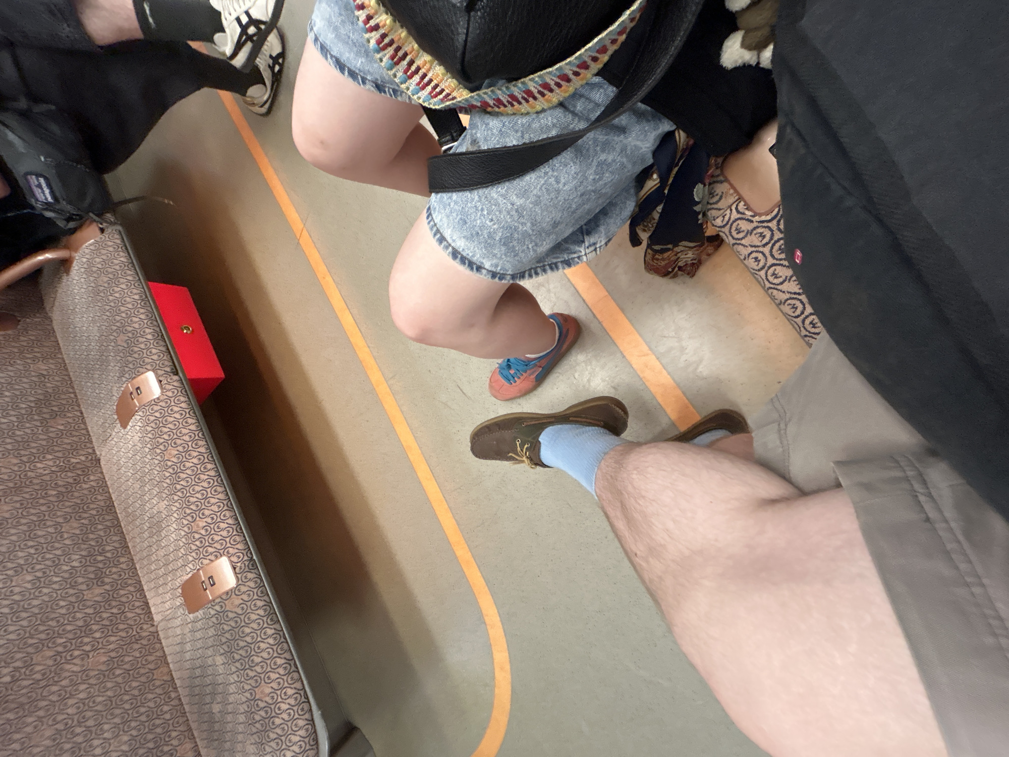 The image shows the interior of a subway car, with the perspective from overhead looking down towards the floor. The car has a grey floor with orange guide lines. A variety of passengers are visible by their lower limbs. To the left is a patterned subway seat with a red square object placed near the backrest. A person wearing black and white sneakers sits adjacent to the seat. In front of the seat, a person wears denim shorts and orange and blue sneakers. To the right, a man in shorts and a grey shirt sits with light blue socks and brown shoes. There is one more person sitting behind the woman in denim shorts. Their feet are adorned with patterned slip-on shoes.
