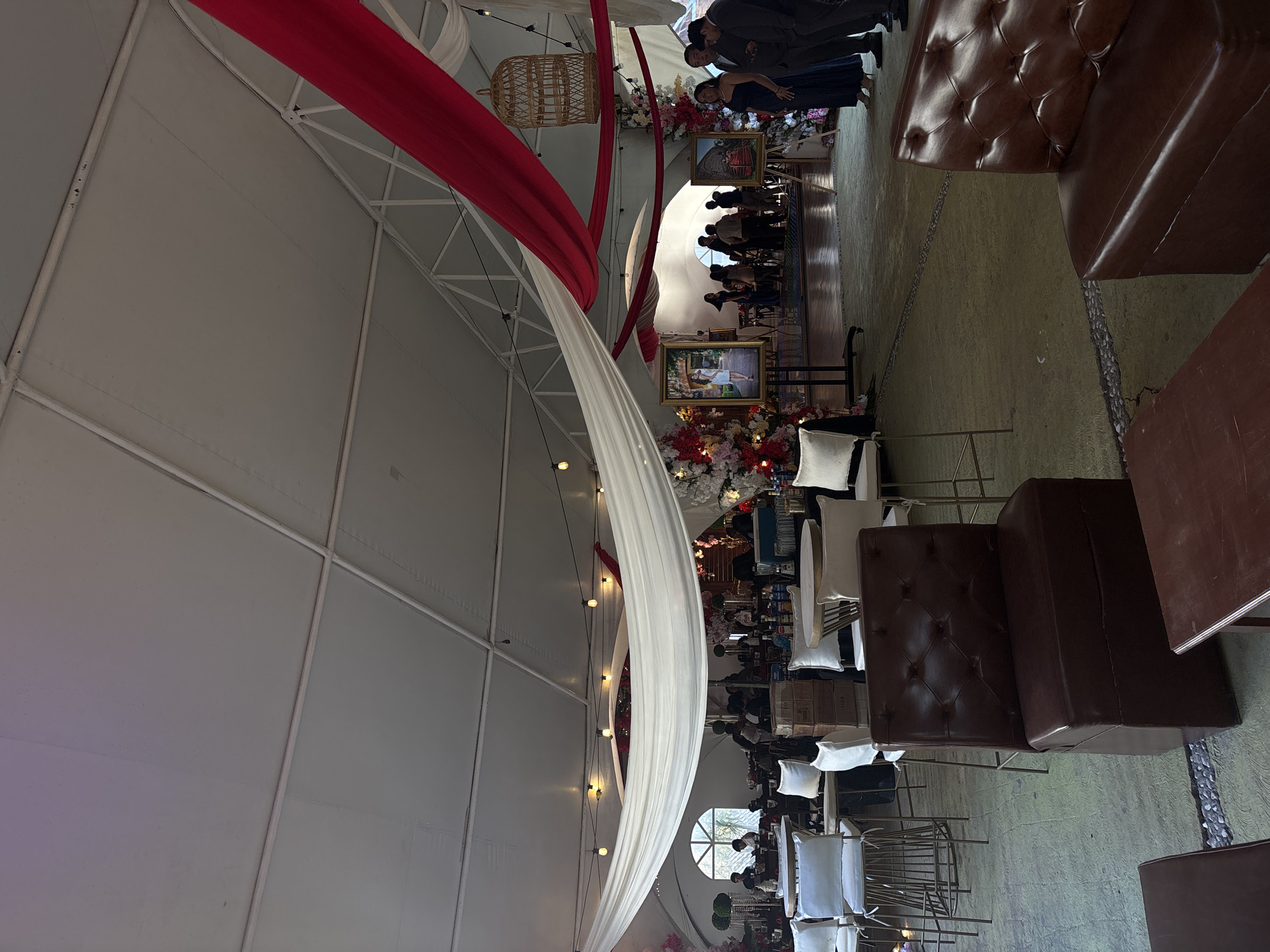 The image depicts an indoor event space, possibly in Chilpancingo, Mexico. The space features a high, white tent-like structure with red and white fabric draping from the ceiling. Hanging lights are strung along the draping. The setup includes seating areas with brown, tufted leather chairs and tables. People are present, some standing near the entrance, while others are seated at tables in the middle. The decor includes floral arrangements and framed artwork. The environment appears to be set for a special occasion or celebration.